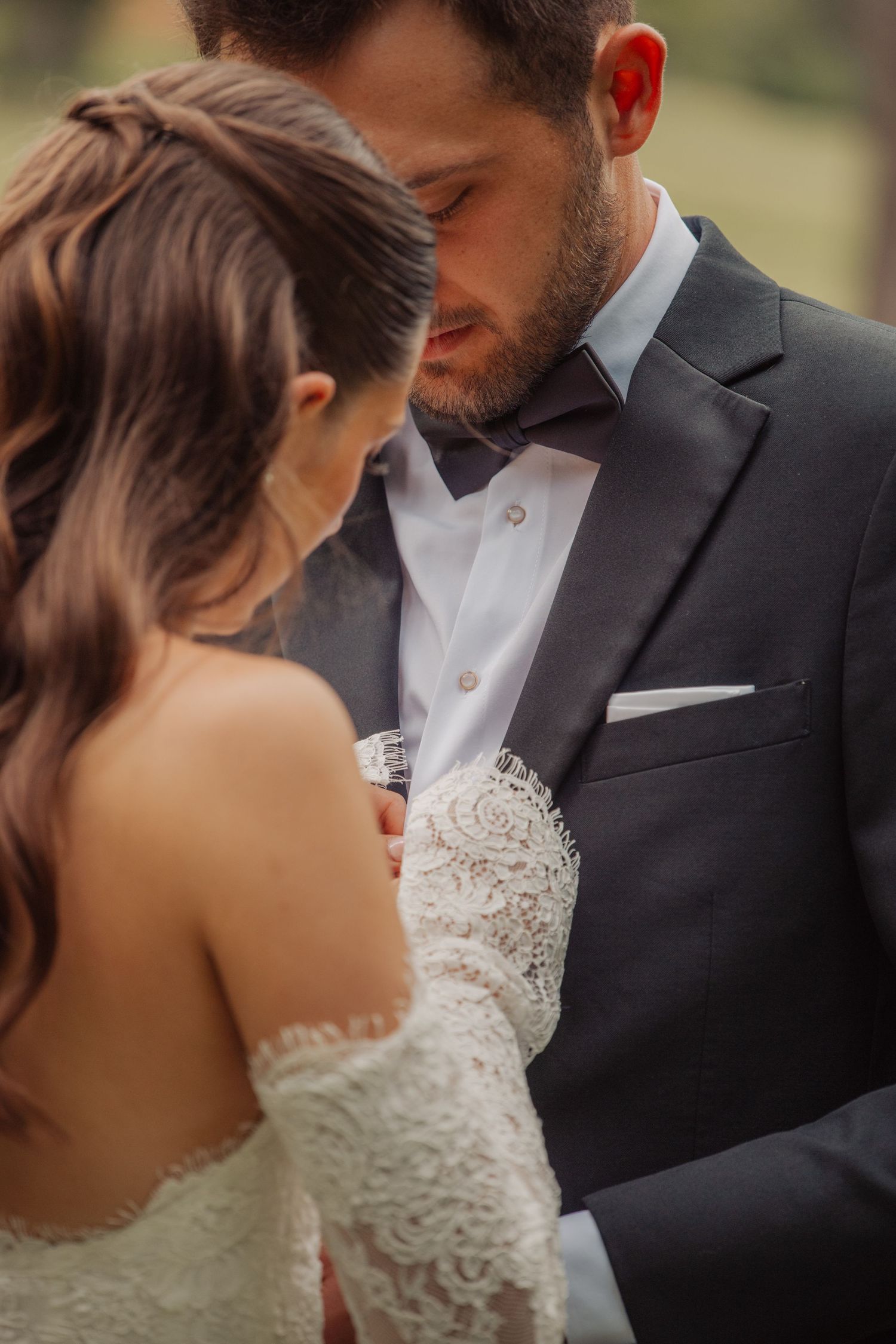 Bride in lace off-shoulder gown and groom in dark suit with bow tie share an intimate moment on their wedding day.