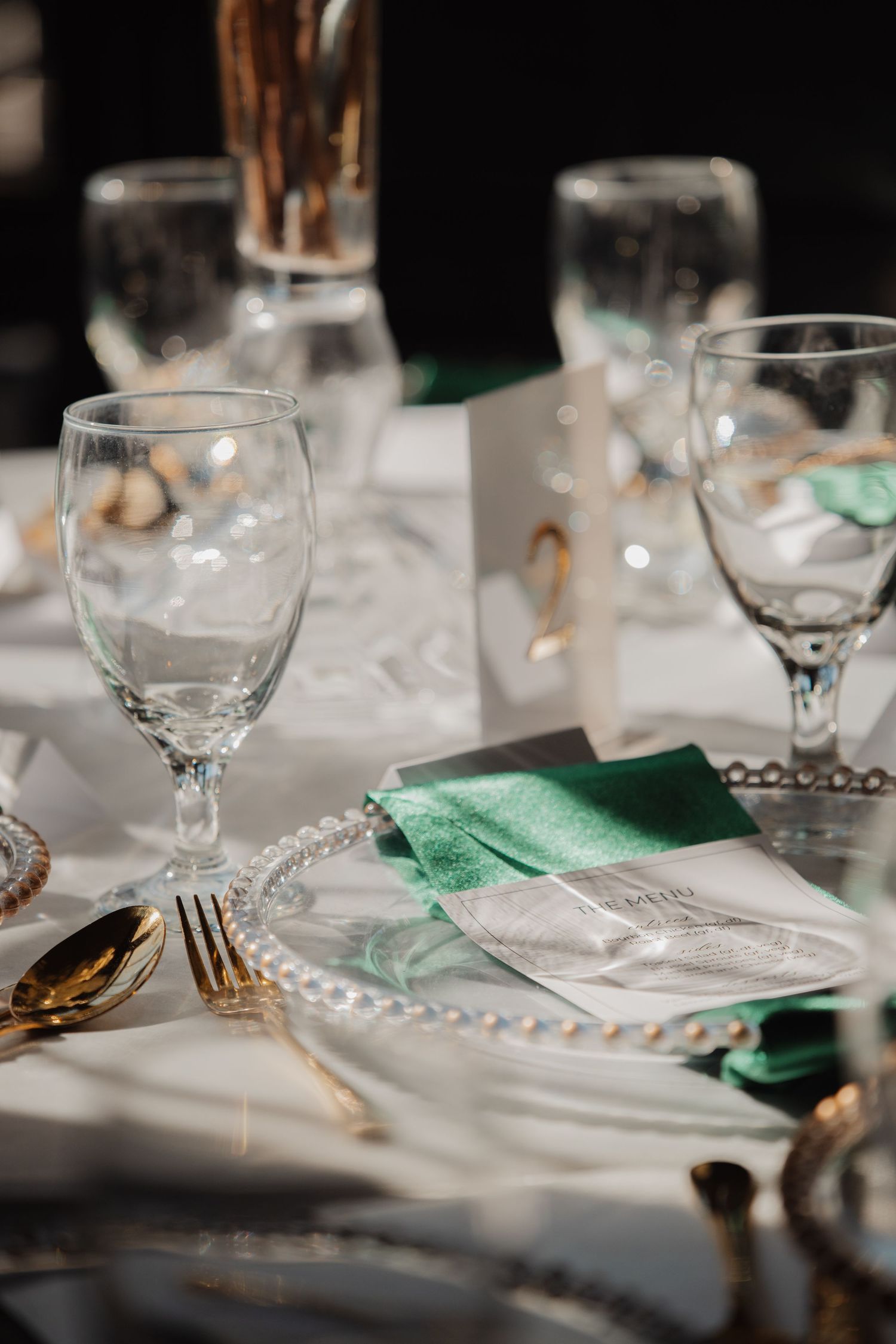 Wedding reception place setting with beaded charger plate, gold cutlery, and emerald green napkin.