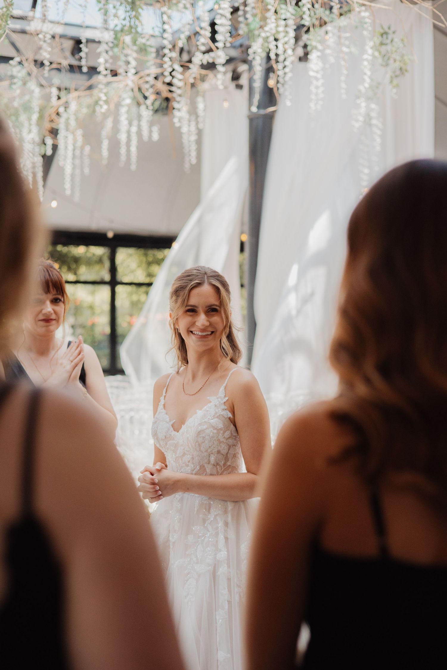 Smiling bride in floral lace gown stands with bridesmaids under hanging greenery in bright indoor venue.