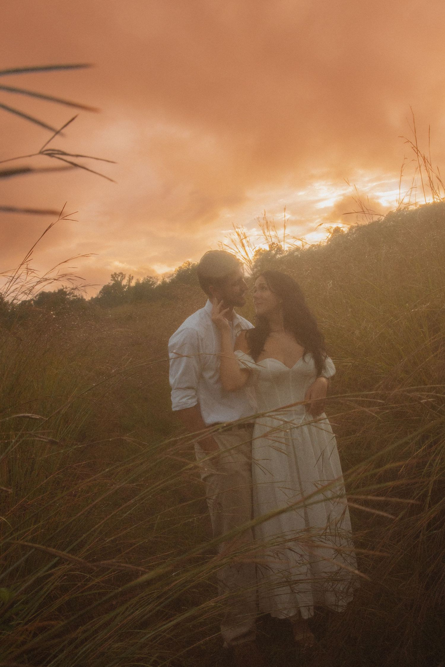 Couple in white embracing in tall grass during a warm golden sunset.