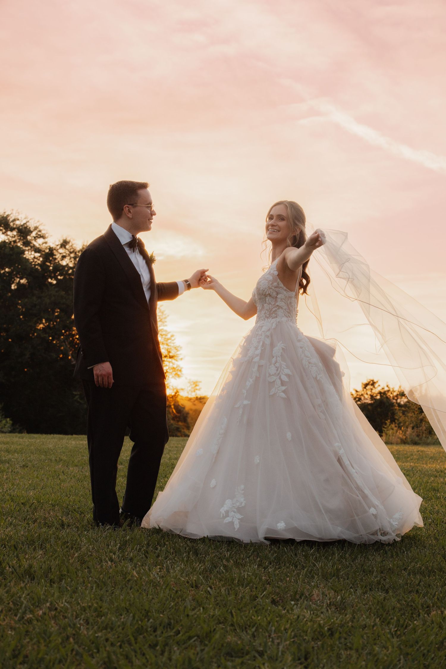 Bride and groom dancing together at golden sunset, bride's veil flowing in a lush green field.