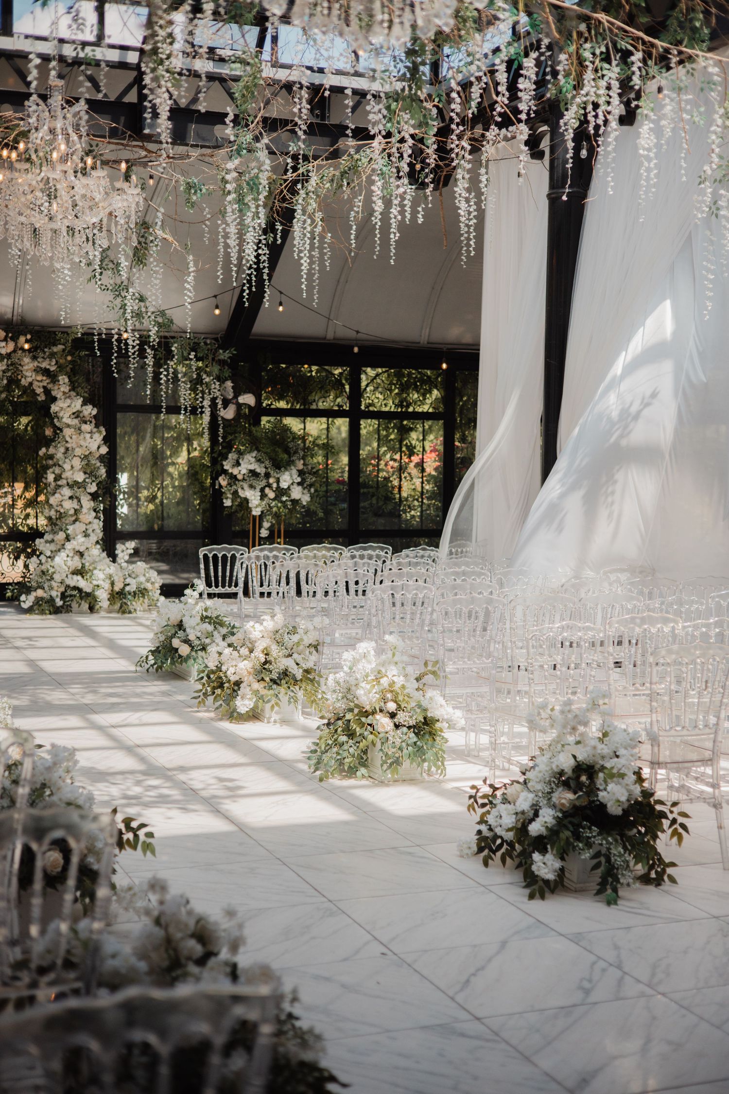 Elegant indoor wedding ceremony venue with white floral aisle arrangements, ghost chairs, and hanging wisteria decor.
