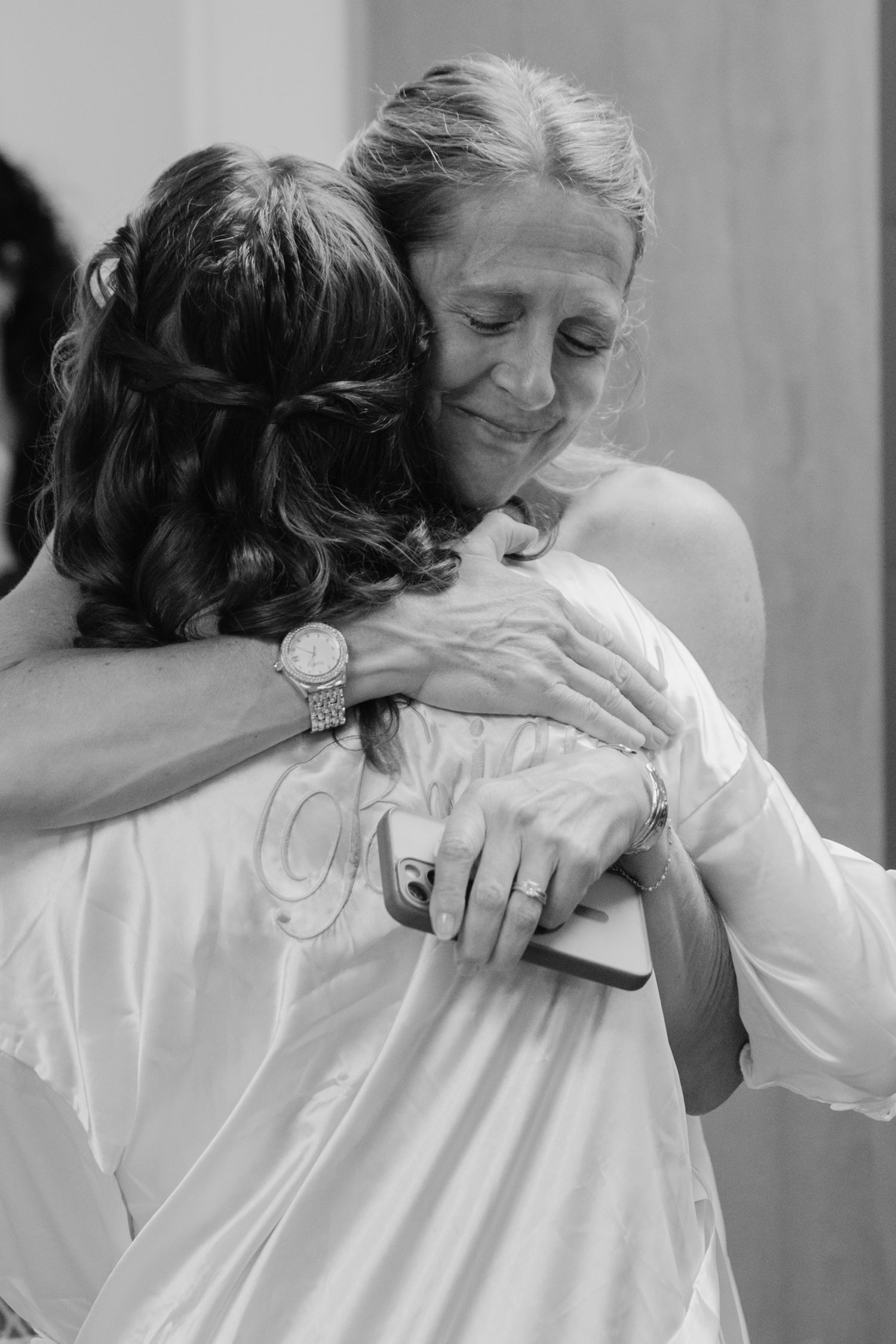 Two women share a heartfelt embrace at a wedding, one in a white bridal robe, captured in black and white.