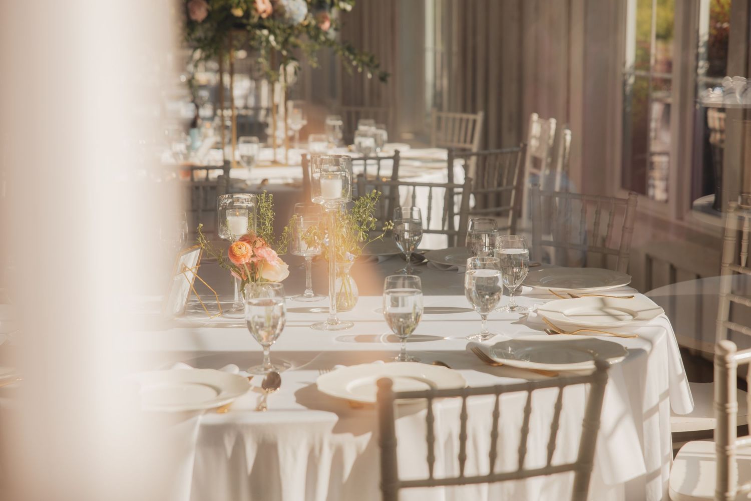 Sunlit elegant wedding reception tables with white linens, gold chiavari chairs, and floral centerpieces.