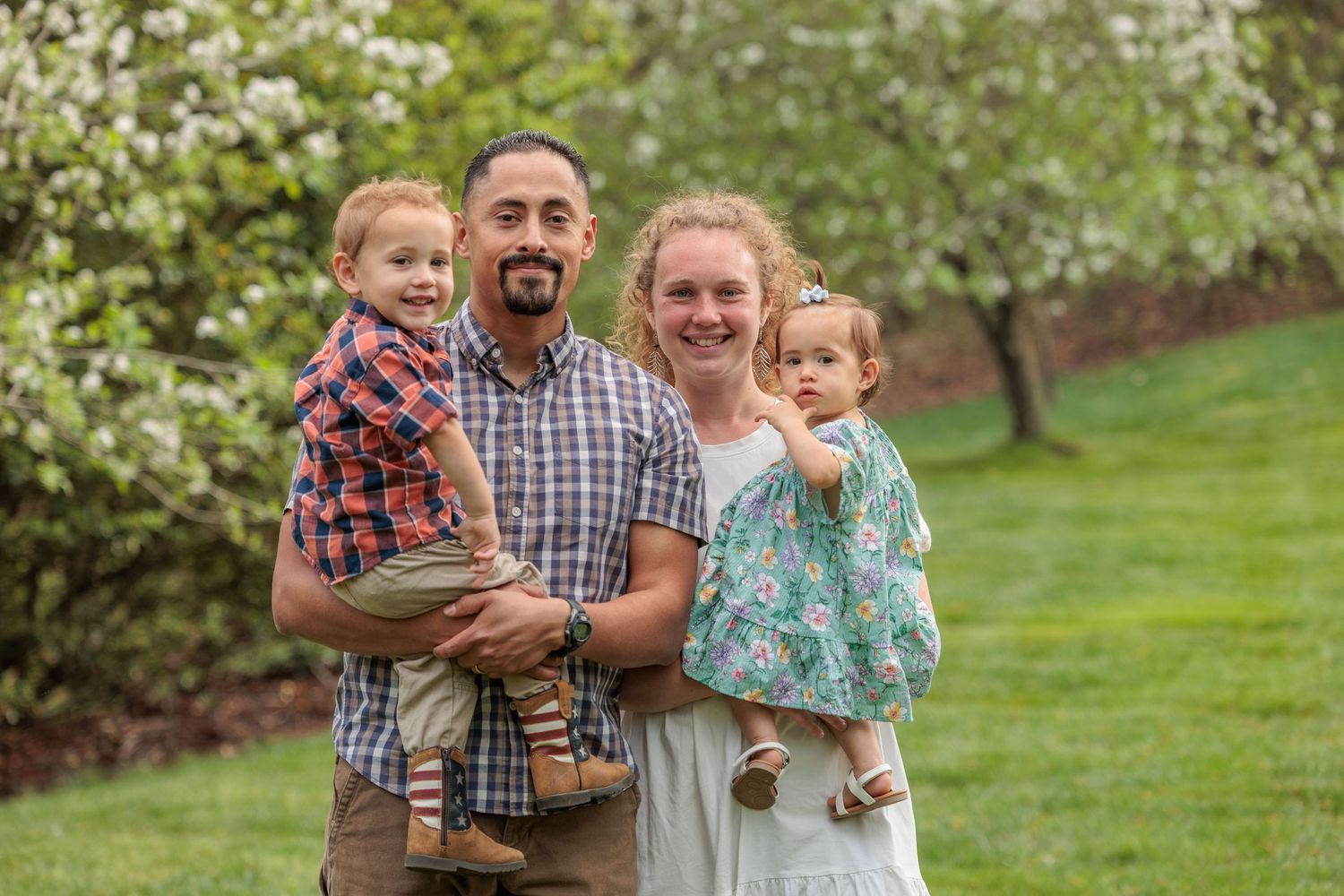 A happy family of four poses outdoors among blooming trees, with parents each holding a young child in spring.
