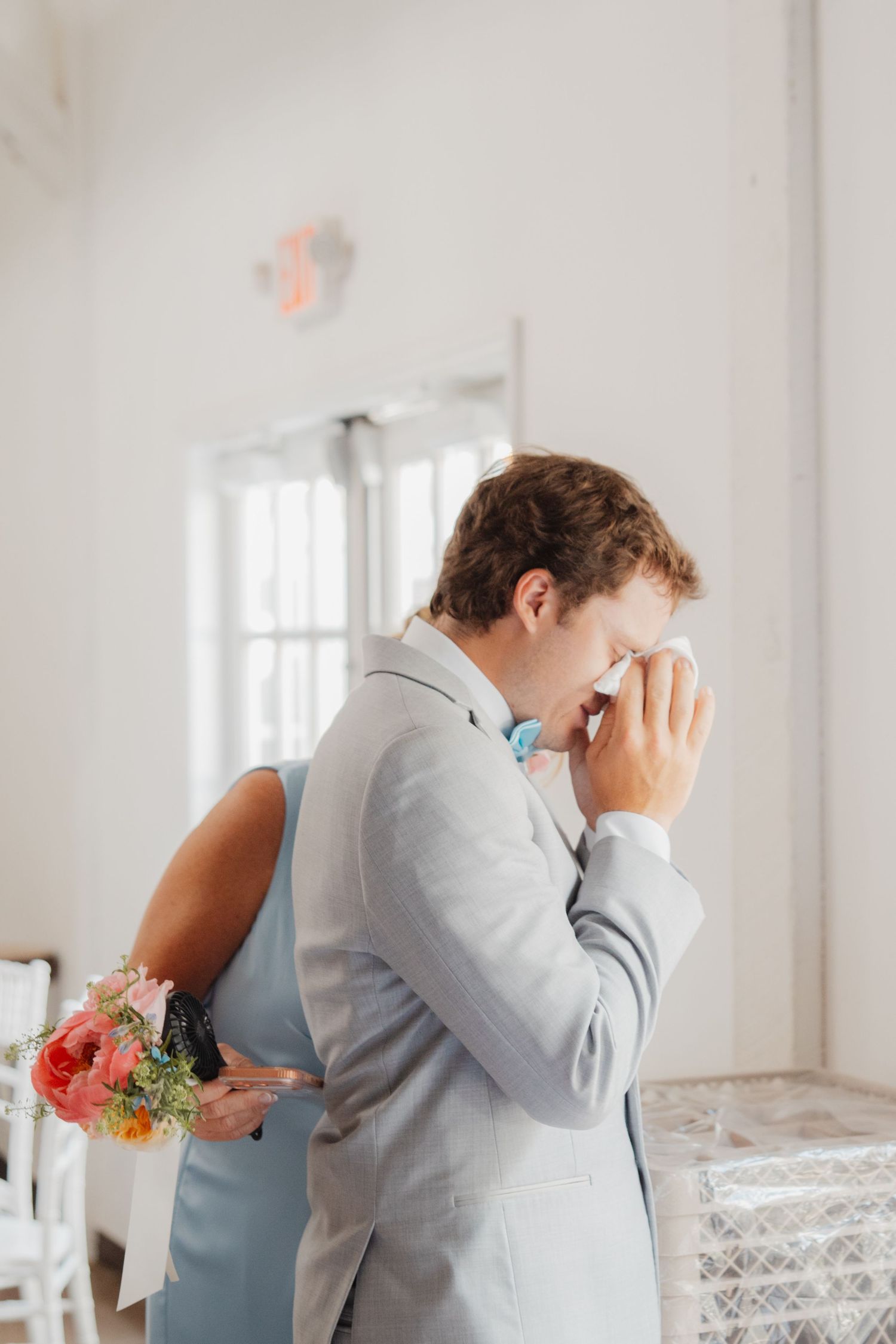 Emotional groom wipes tears while embracing someone holding a colorful bridal bouquet indoors.