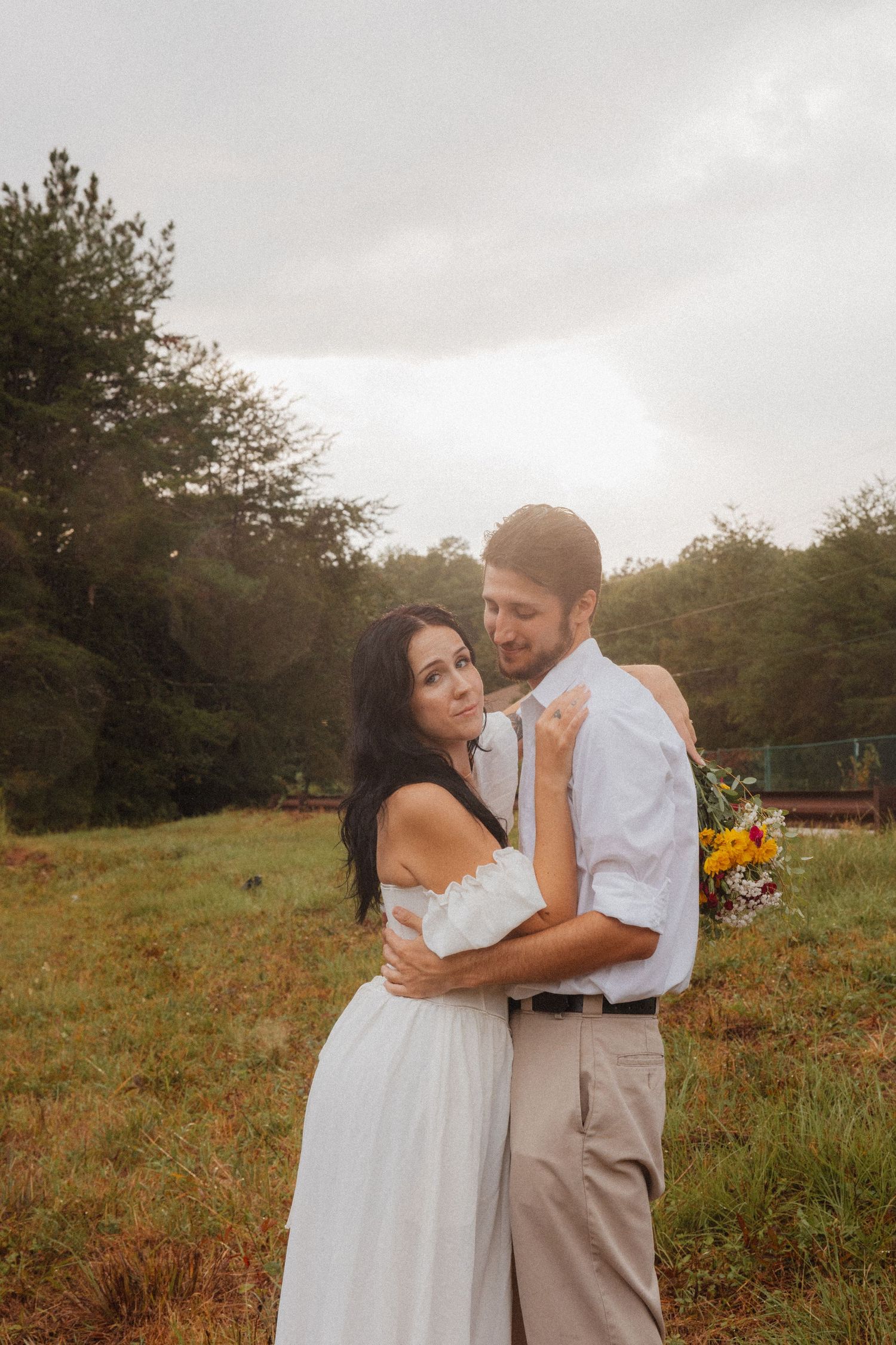 Couple embracing in a green meadow, woman in white dress holding wildflower bouquet.