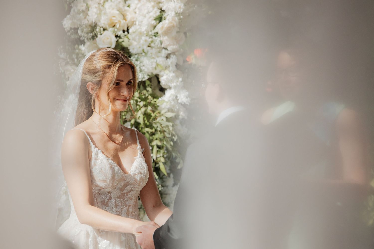 Radiant bride smiling in lace wedding dress before white floral arch during wedding ceremony.
