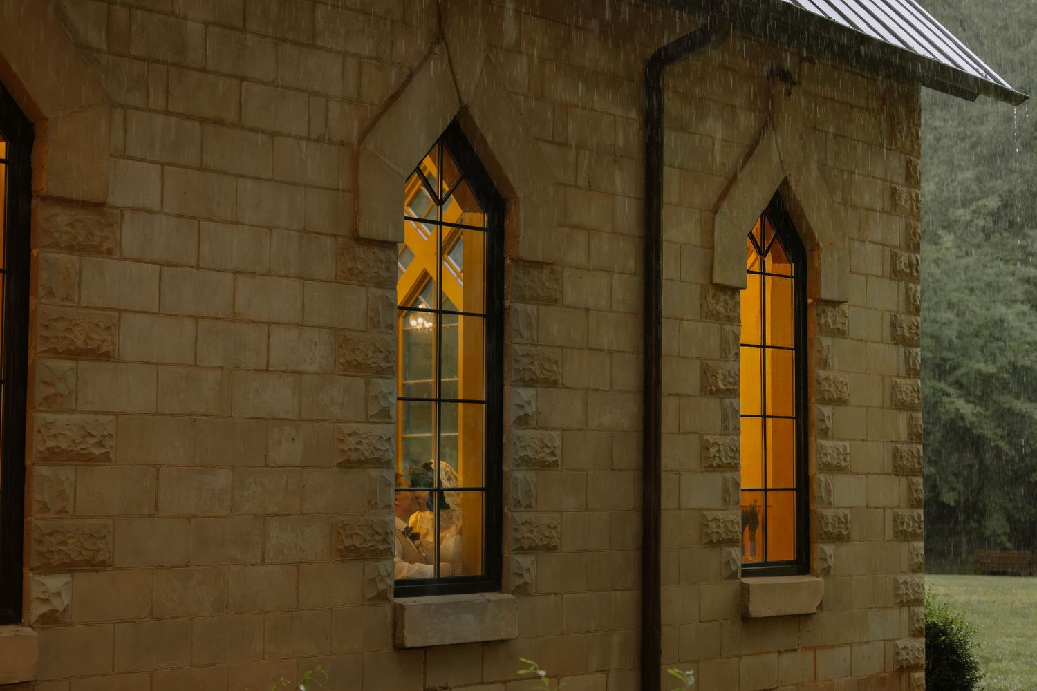 Stone church exterior at dusk with glowing arched stained glass windows illuminated by warm golden light.
