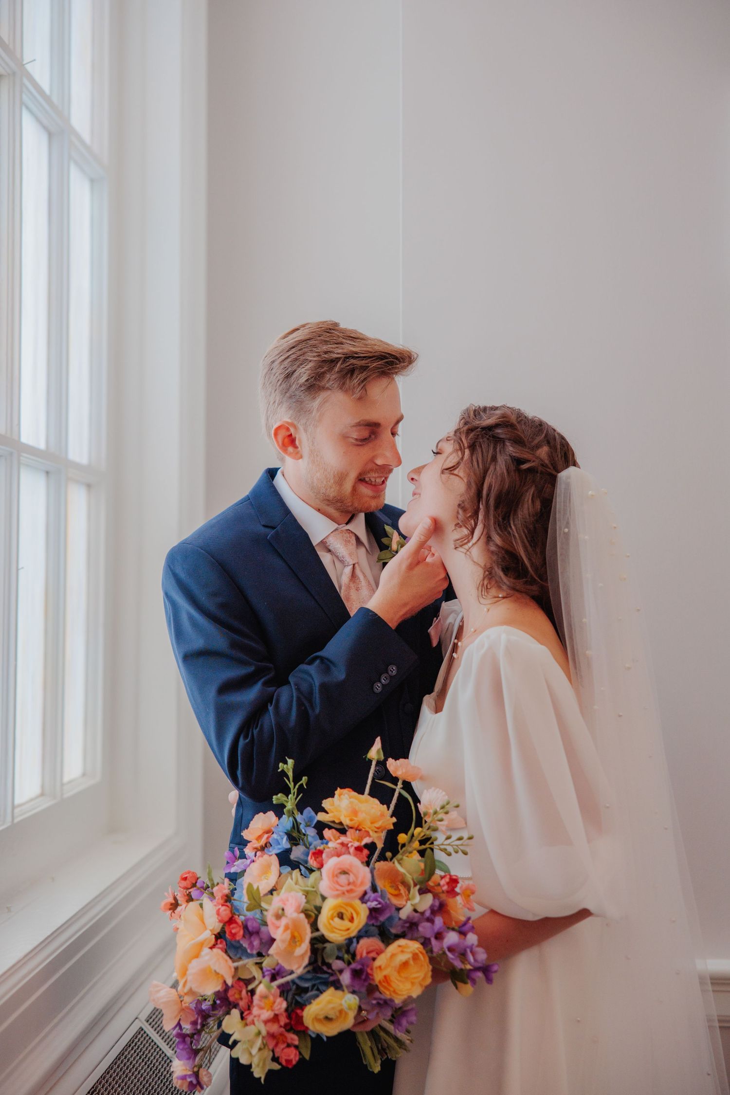 A groom in a navy suit tenderly touches his bride's face as she holds a vibrant colorful bouquet.