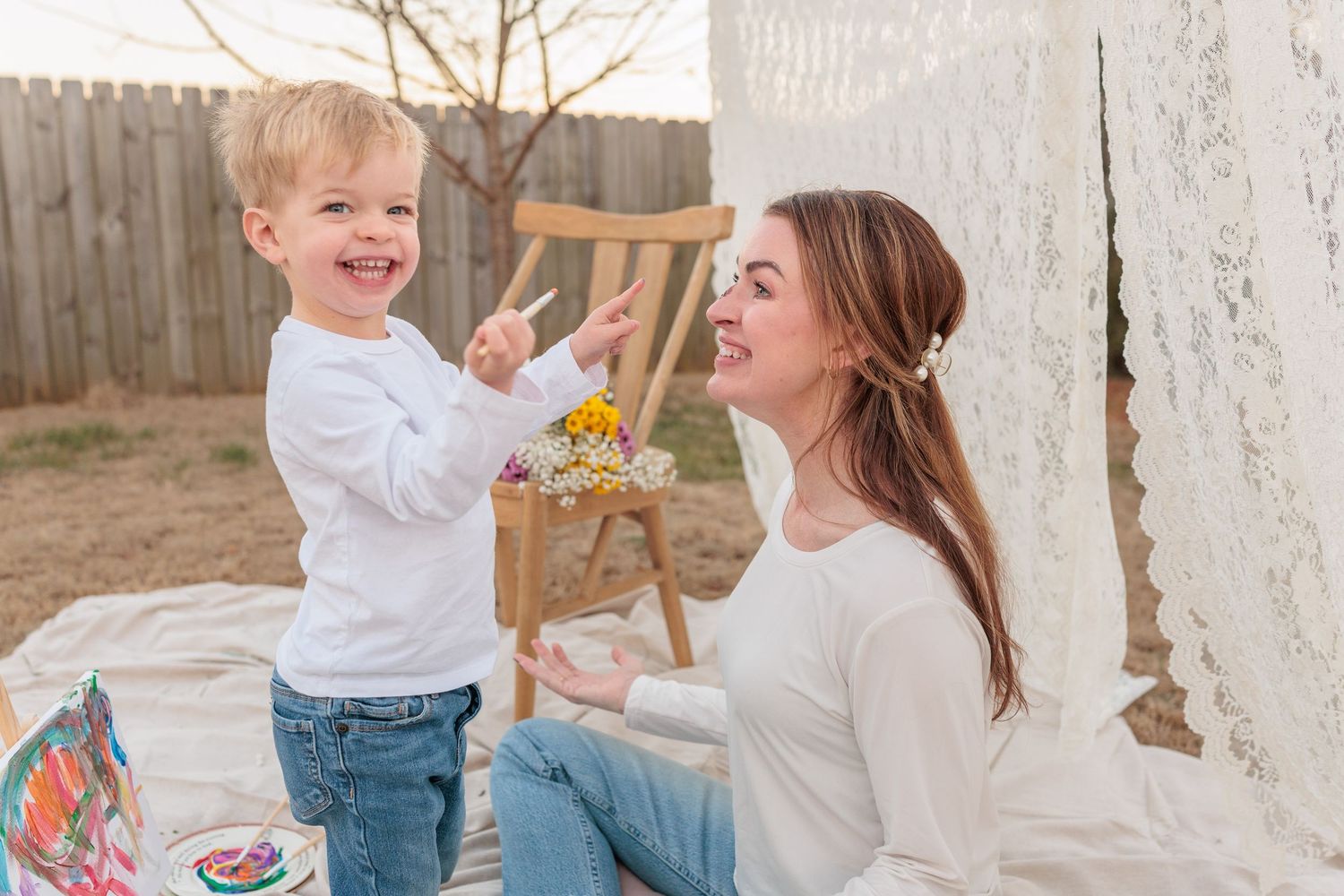 A young toddler boy laughs while painting with a brush alongside a smiling young woman outdoors.