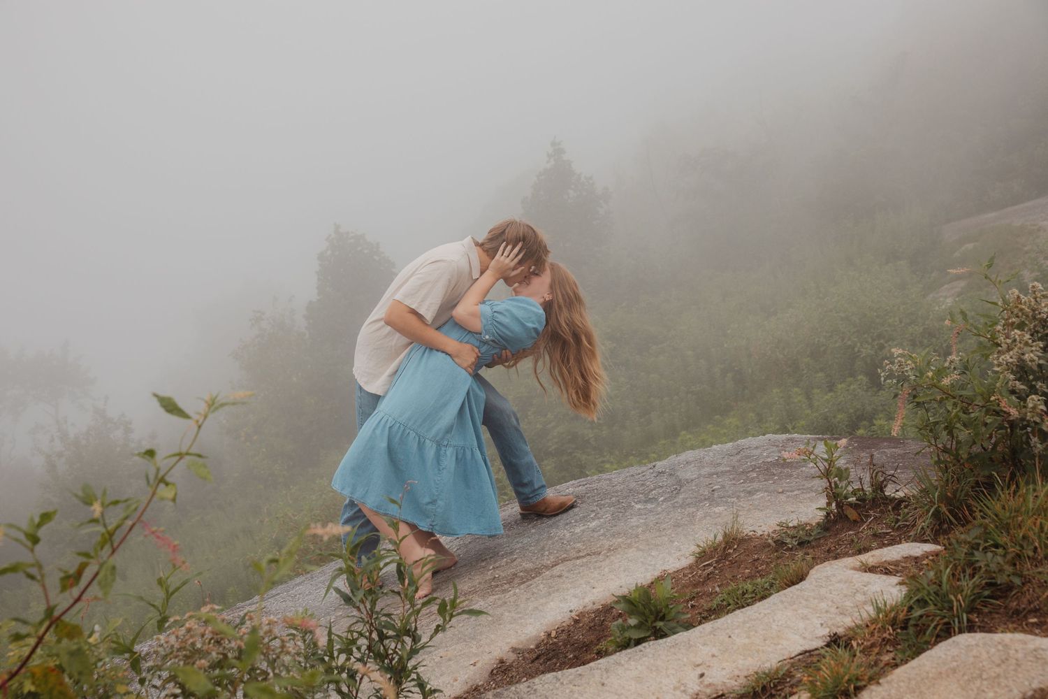 Young couple sharing a passionate dip kiss on a foggy rocky overlook surrounded by green shrubs.