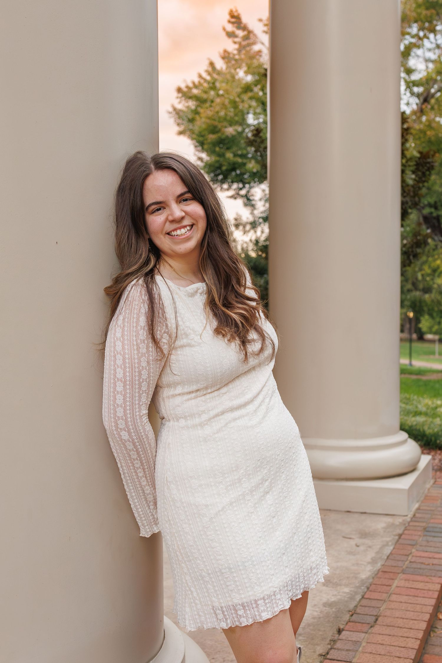 Young woman in white lace long-sleeve mini dress smiling between elegant columns at golden hour.