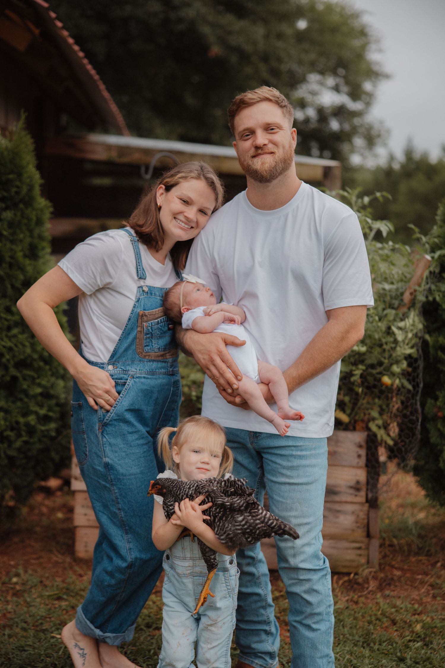 Young family of four poses outdoors on farm, parents in white tees and denim, toddler holding black hen, dad cradling newborn.