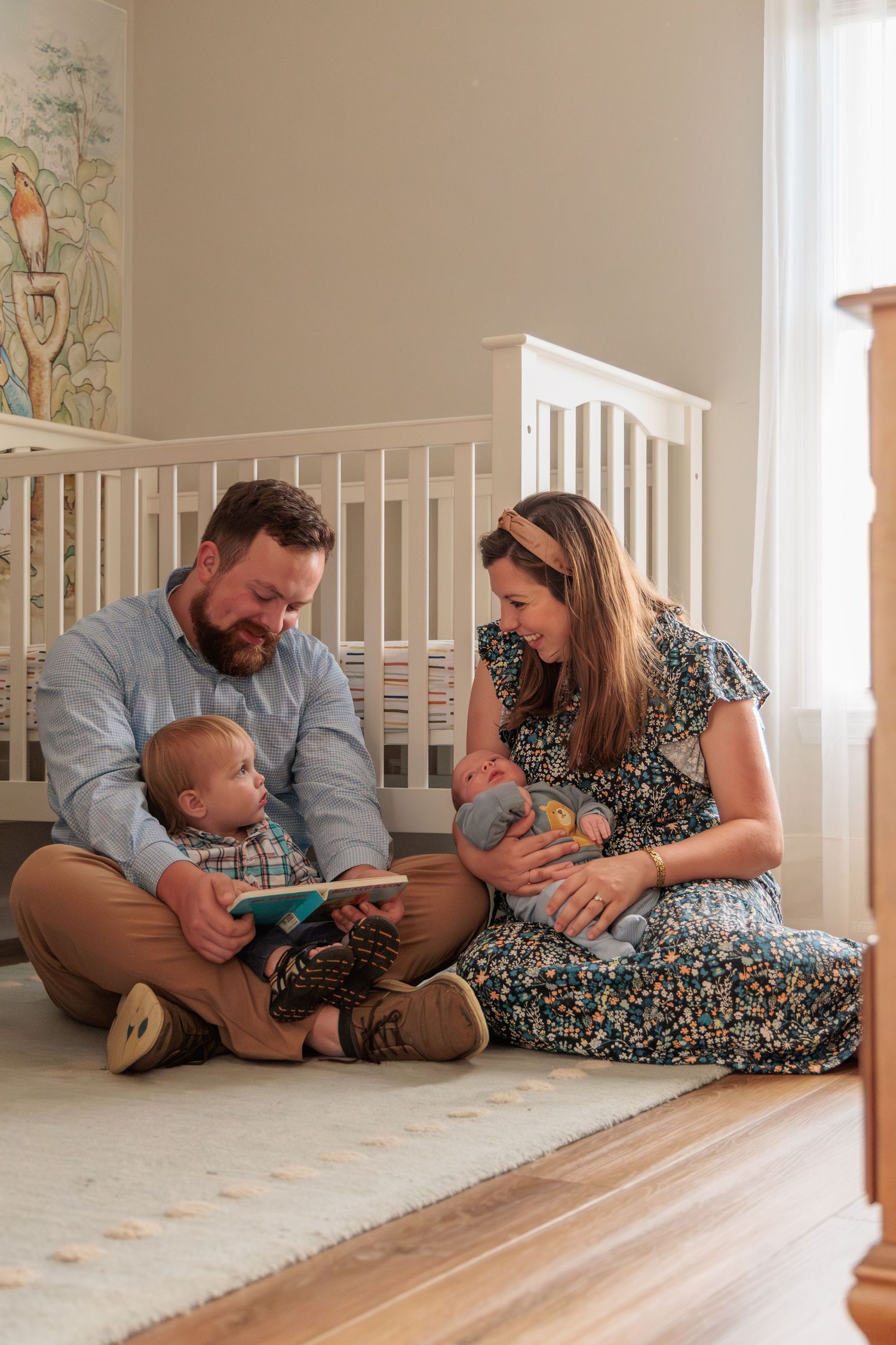 Young family sitting on nursery floor, father reading to baby while mother holds infant near white crib.