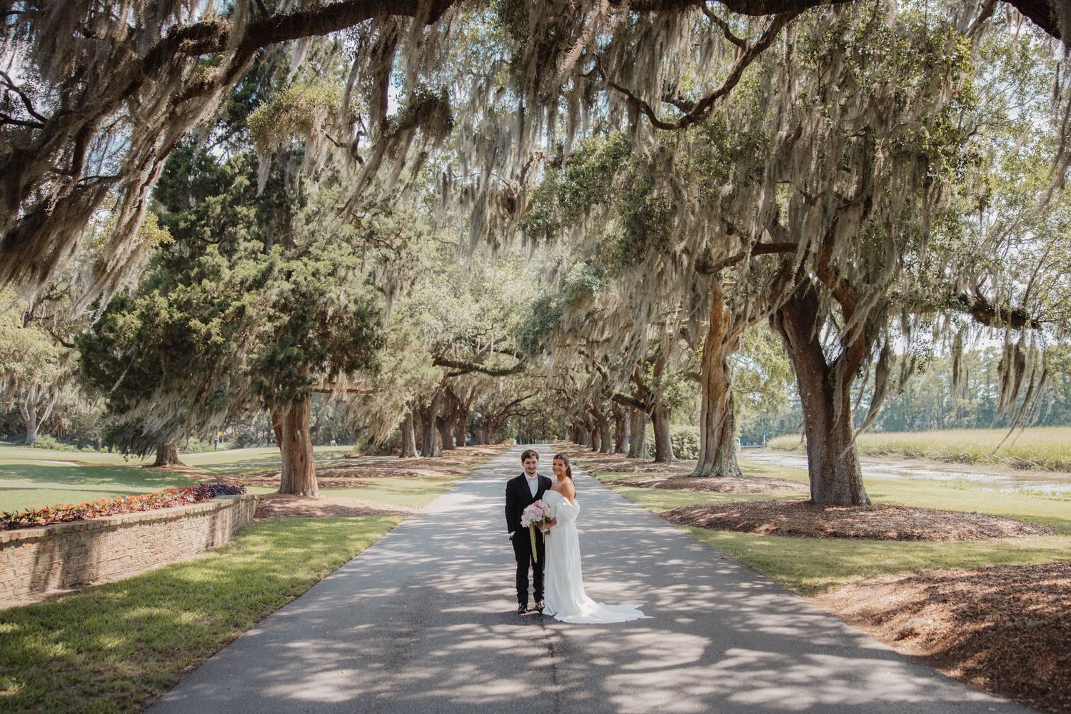 Bride and groom walking together under stunning canopy of moss-draped oak trees on scenic path.