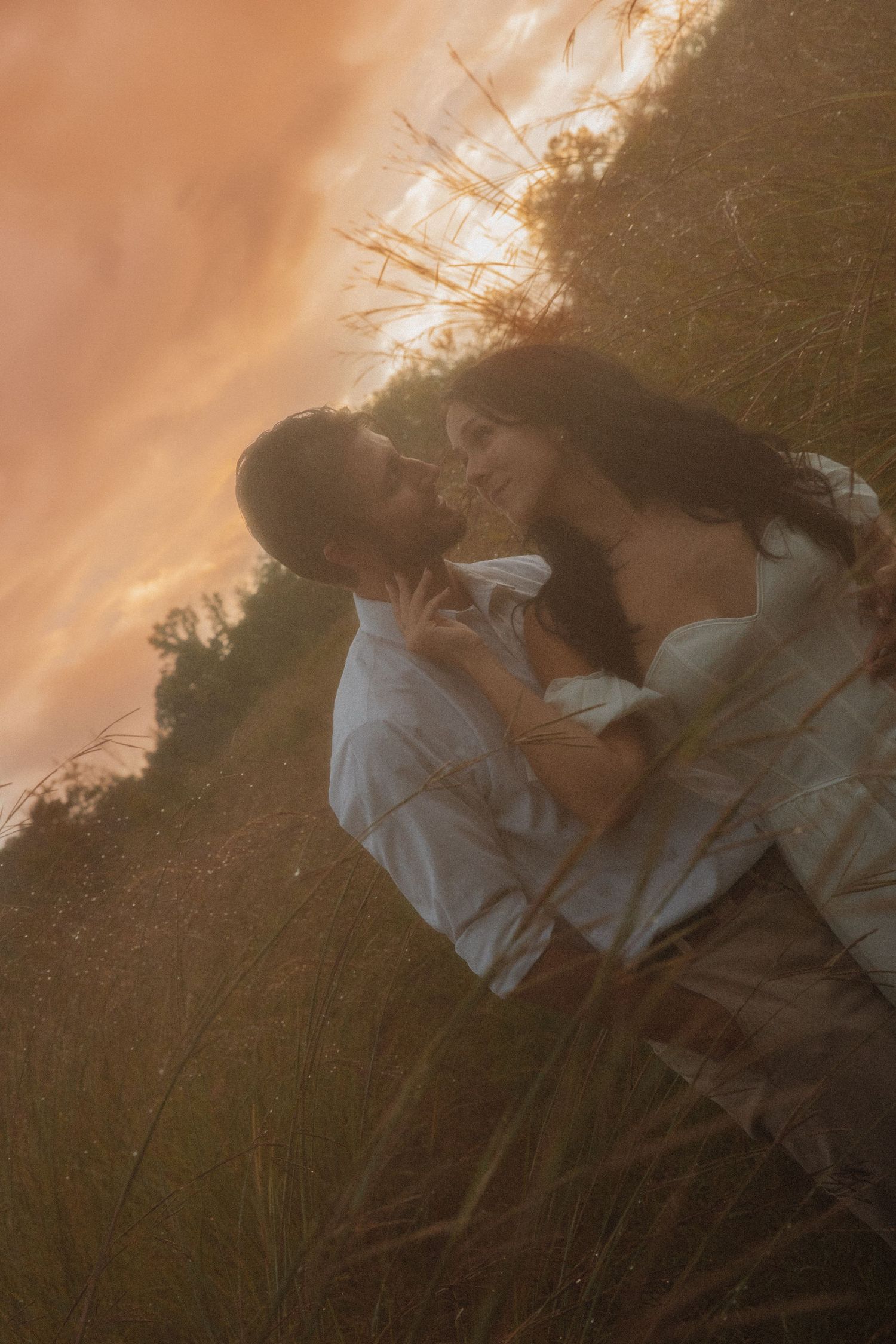 Couple leaning in for a kiss in tall grass field bathed in warm golden sunset light.