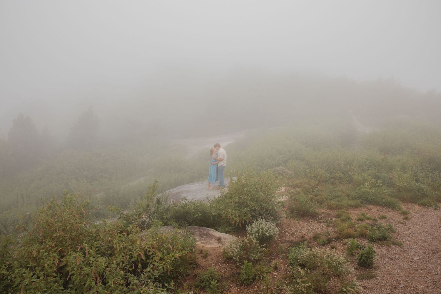 Young couple embracing on a misty mountain path surrounded by lush green shrubs and fog.