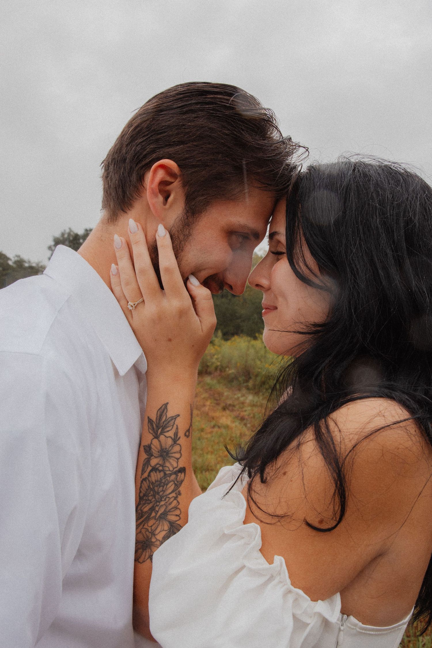 Intimate close-up of couple touching foreheads outdoors, woman with floral arm tattoo and ring.