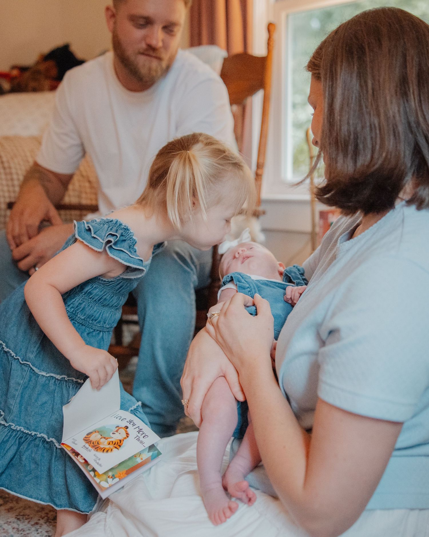 A young family sits together indoors as a toddler girl in blue reads a book beside her mother holding a newborn baby.