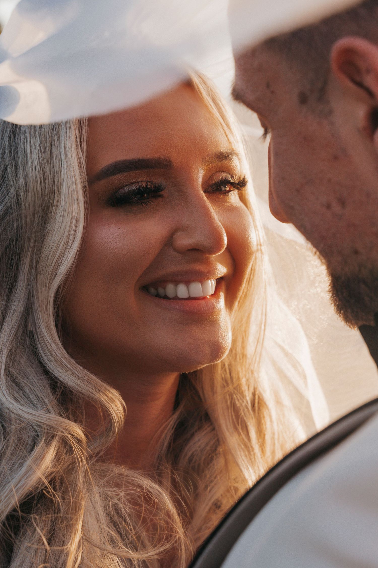 A close-up of a smiling bride with wavy blonde hair and her groom, glowing in warm golden light.