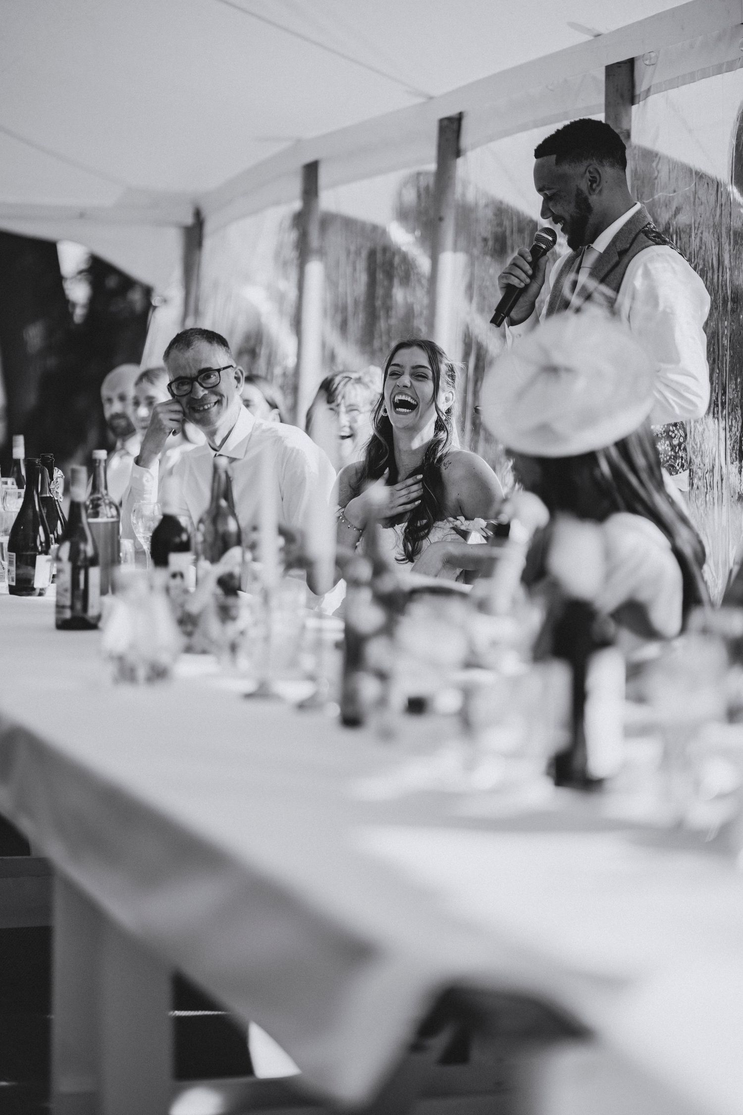 Black and white photo of a best man giving a speech into a microphone as the bride laughs at a reception table.