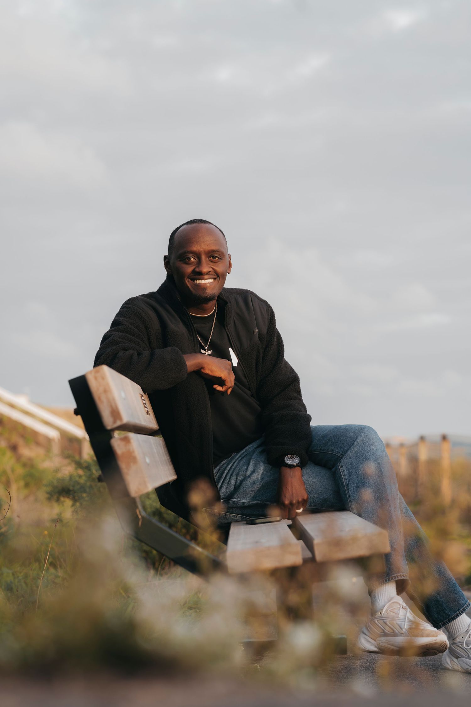 Smiling man in a black jacket and jeans sits on a wooden bench surrounded by grass and dunes under a soft cloudy sky.