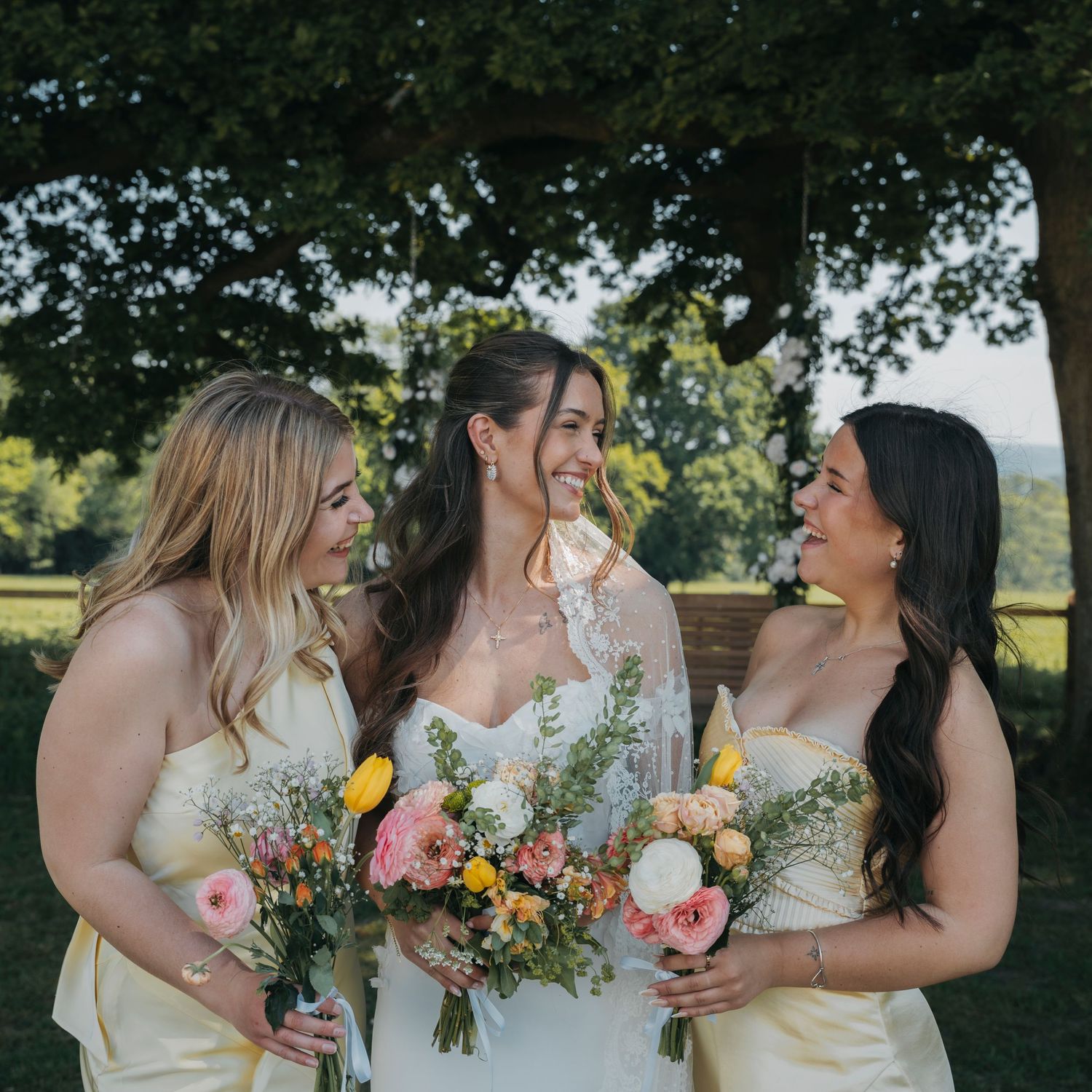 Bride and two bridesmaids in yellow dresses hold pink and white bouquets and smile together under a large tree.