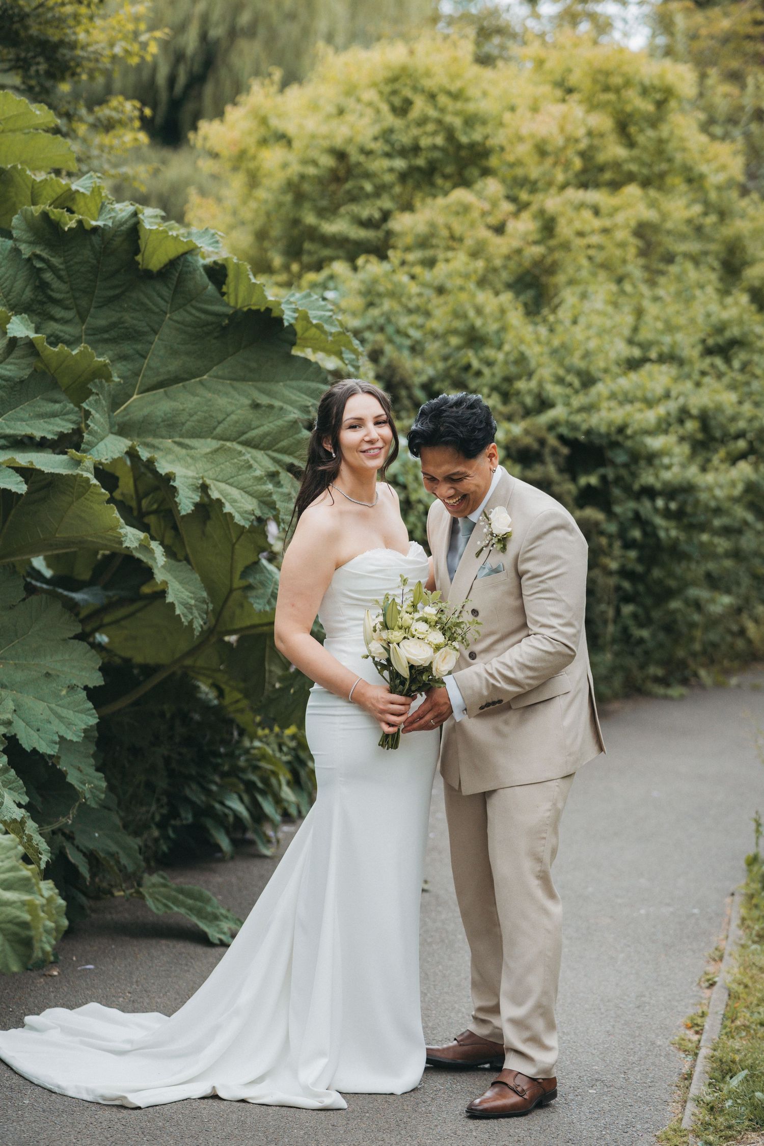Smiling bride in a strapless white gown holds a bouquet beside her groom in a tan suit on a garden path with lush green foliage.