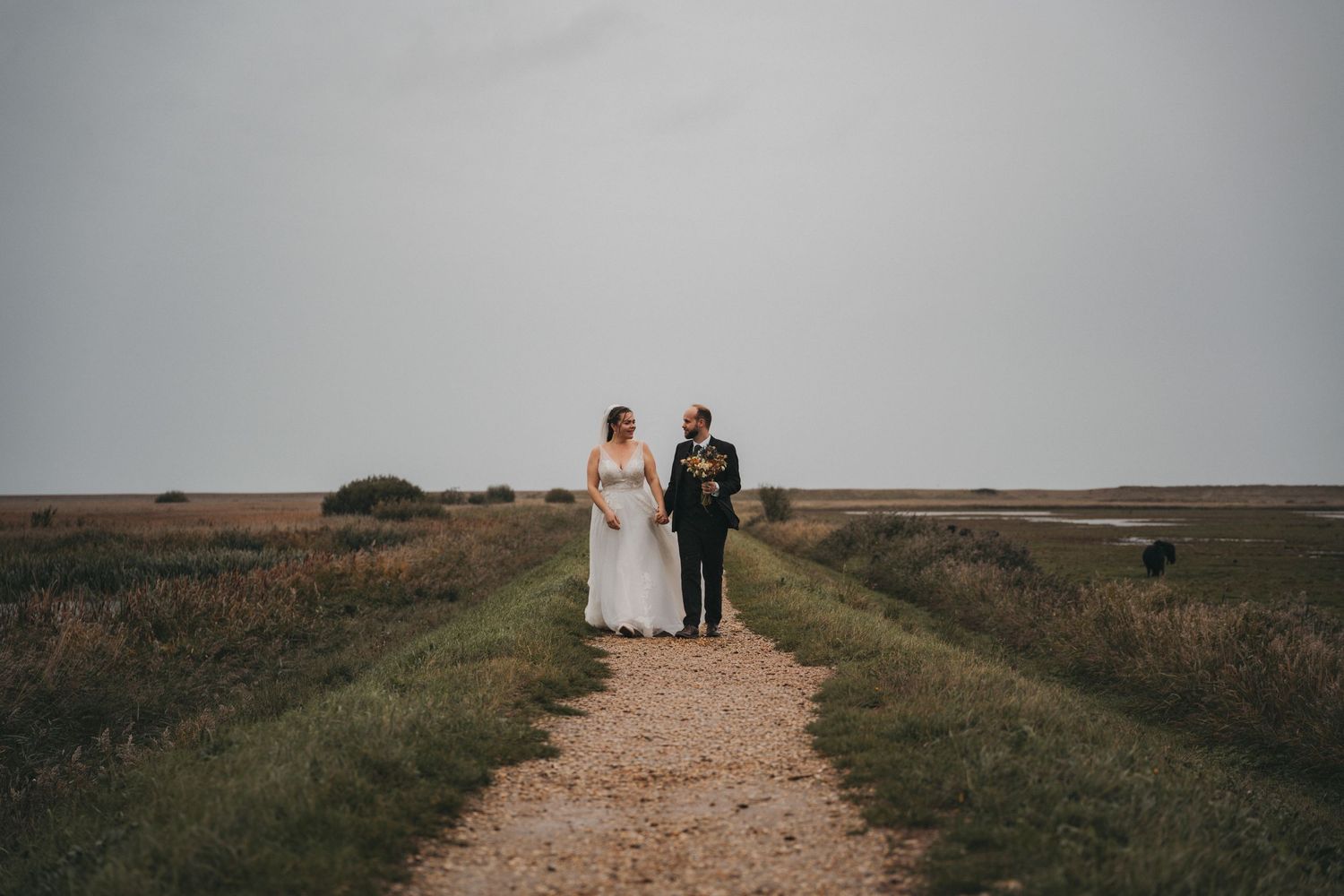 Bride and groom hold hands and walk along a gravel path through windswept marshland under a moody grey sky.