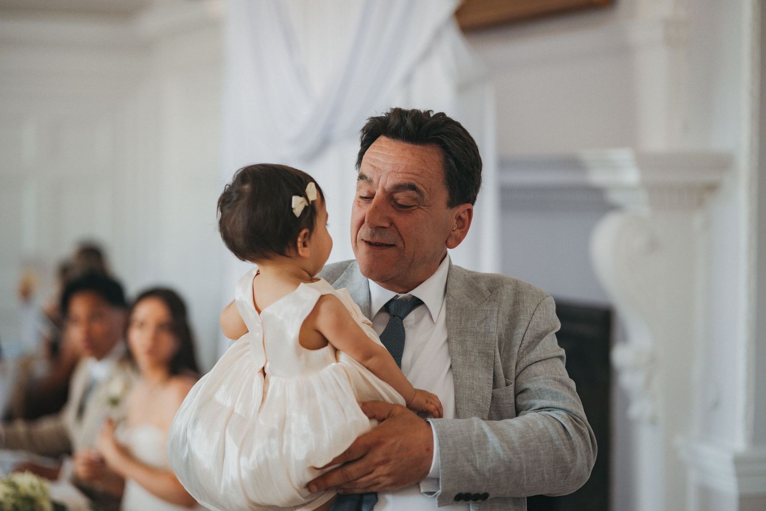 A smiling older man in a gray suit holds a little girl in a white dress during a wedding celebration.