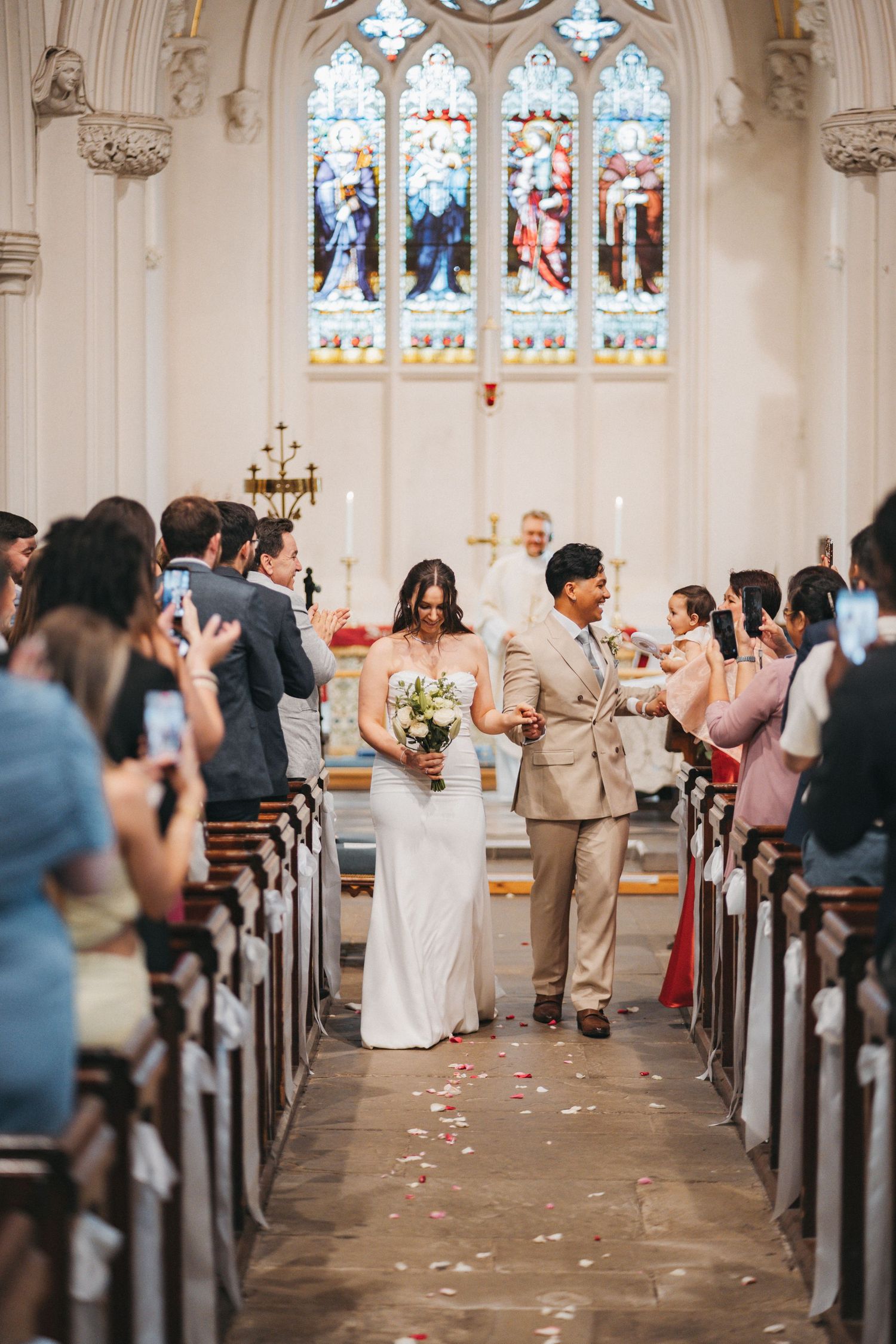 A bride and groom walk hand in hand down the church aisle as guests take photos beneath stained glass windows.