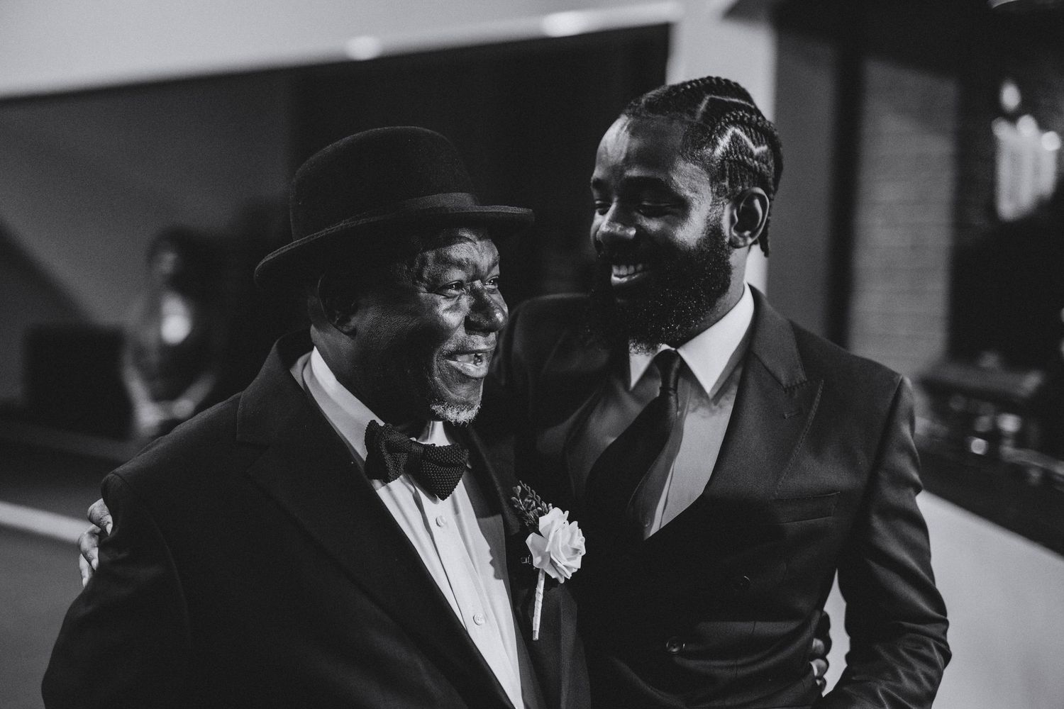Black and white portrait of two smiling men in formal wedding attire, one wearing a hat and bow tie, sharing a joyful moment.