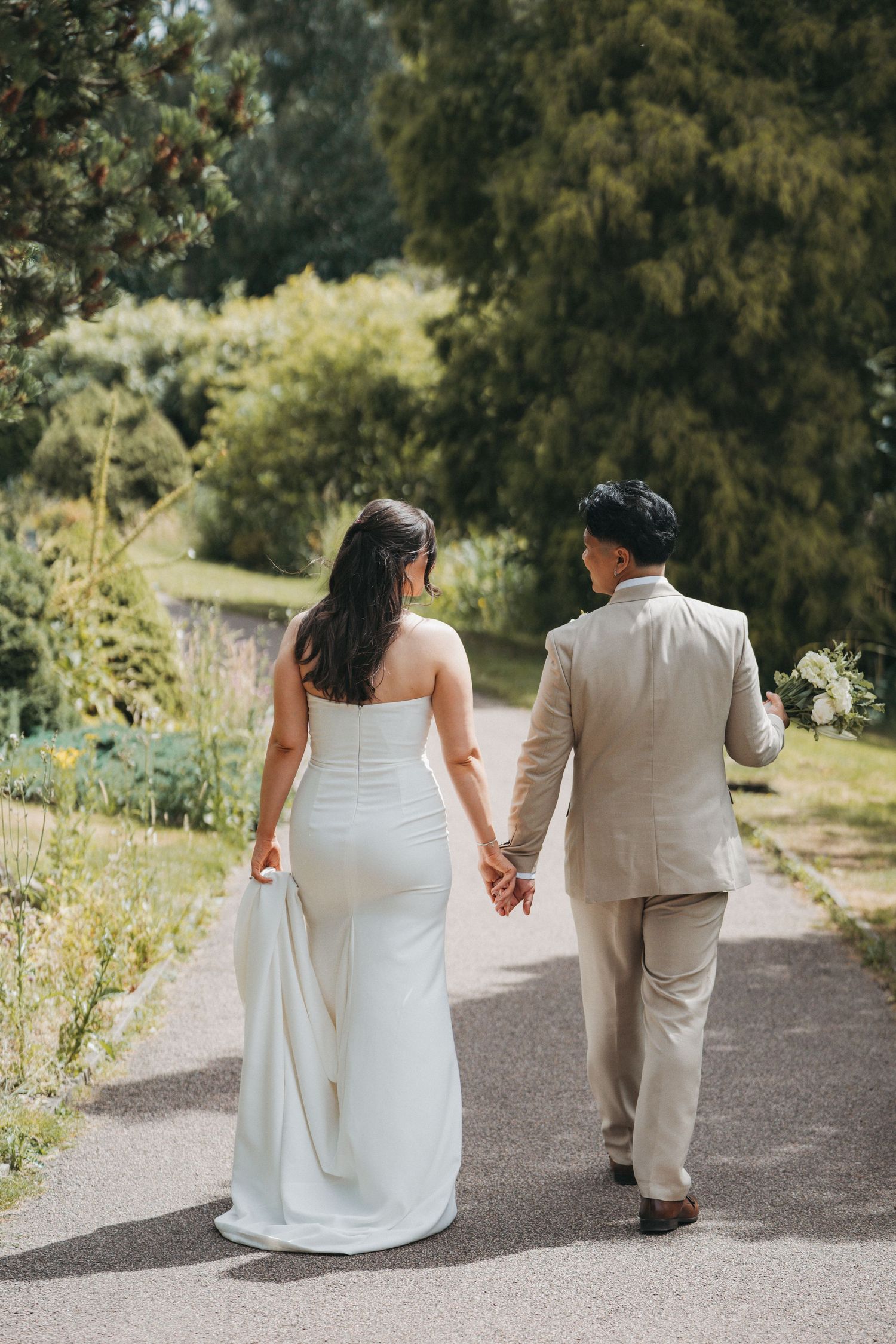 Bride in a white fitted gown and groom in a beige suit walk hand in hand along a sunlit garden path surrounded by trees.