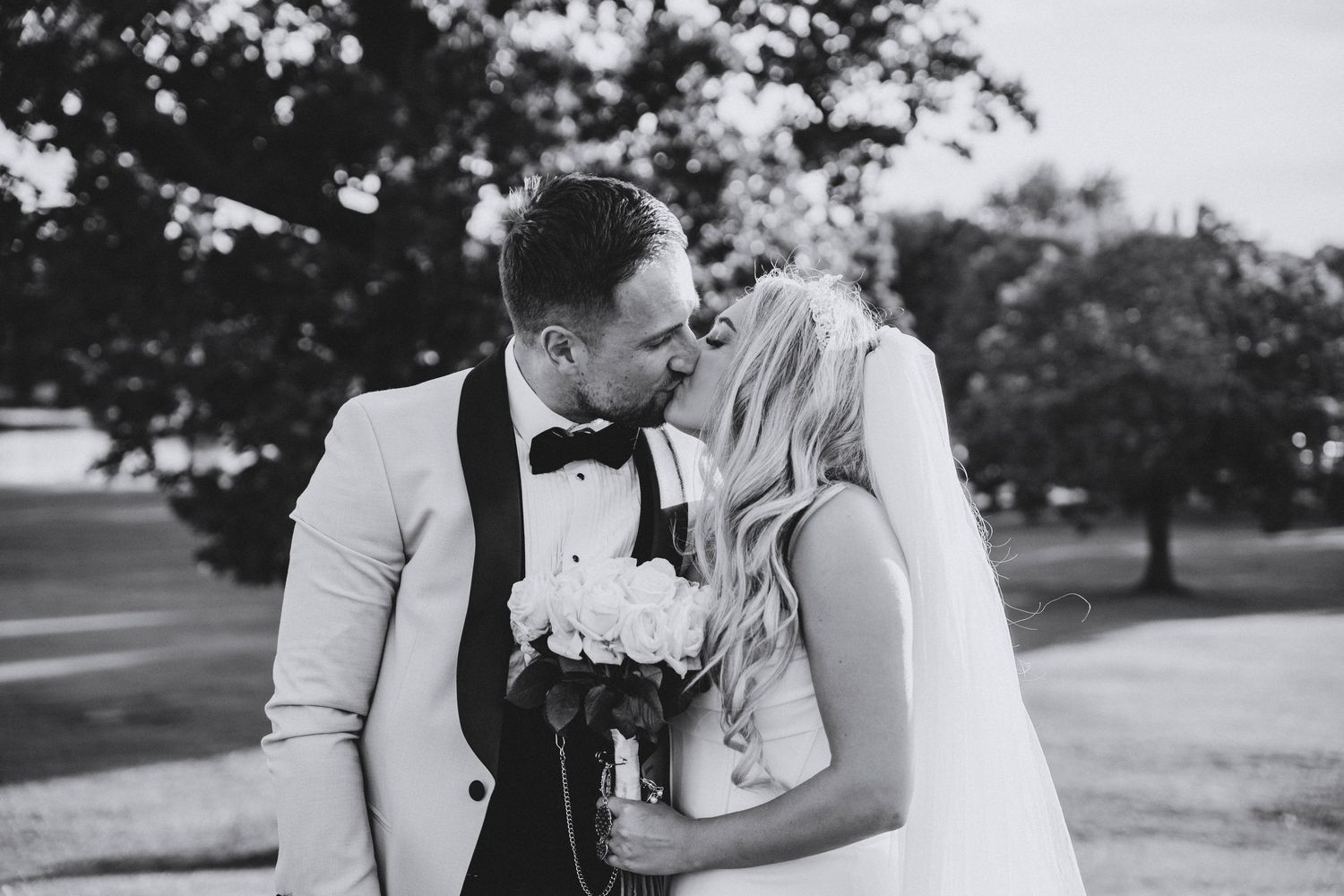 Black and white photo of a groom in a bow tie kissing his bride in a veil as she holds a bouquet outdoors.