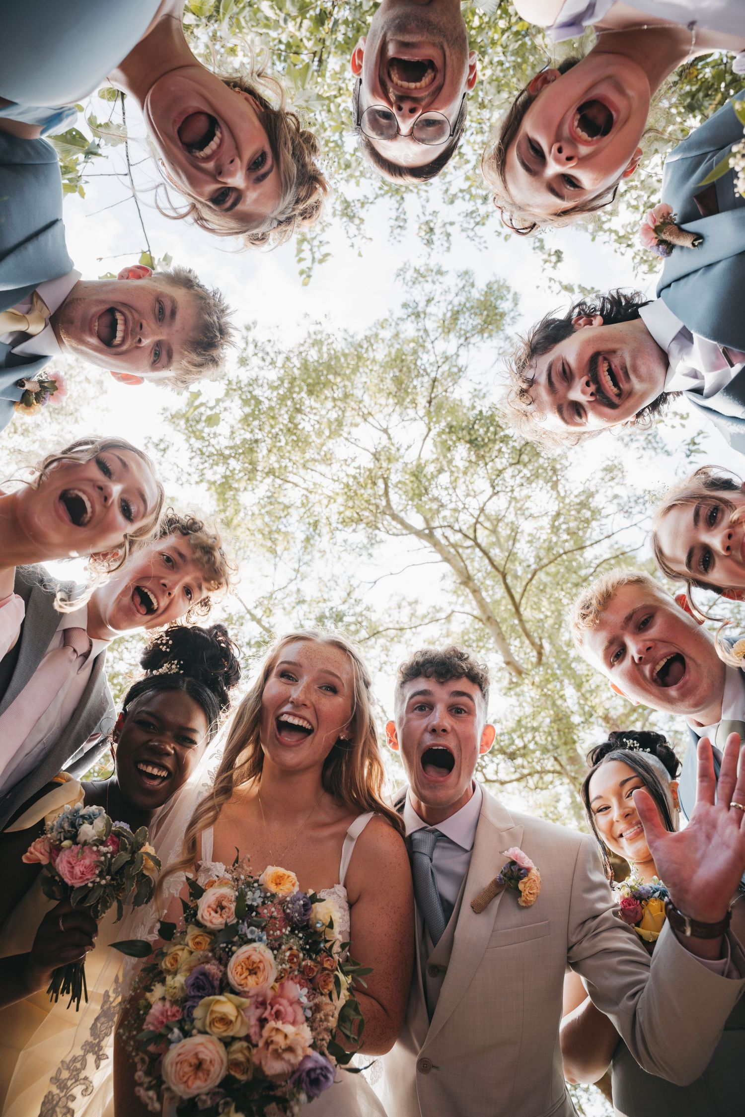 Low-angle view of a laughing bride, groom and wedding party gathered in a circle beneath leafy trees with colorful bouquets.
