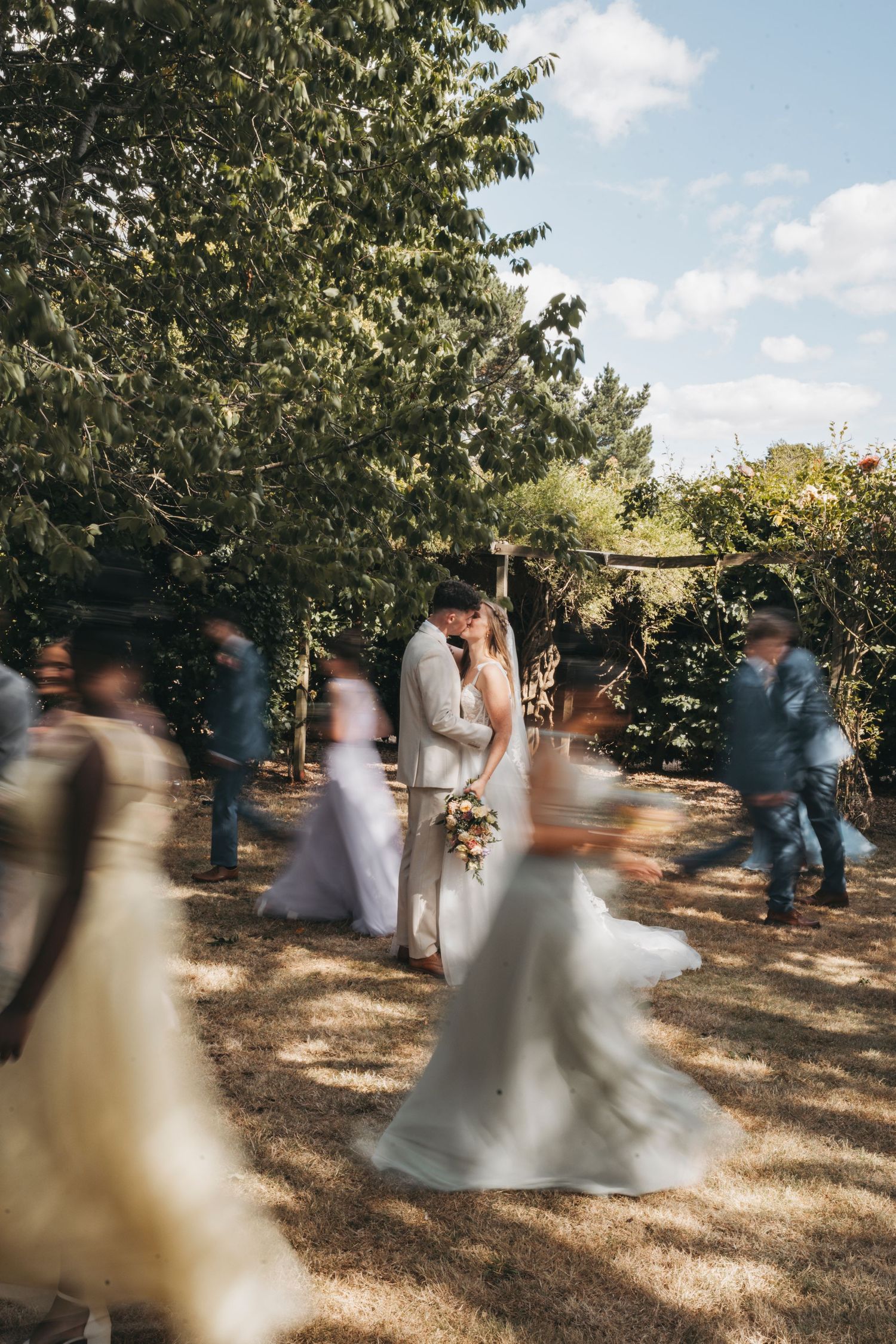 Bride and groom kiss in a sunlit garden while blurred wedding guests in motion move around them.