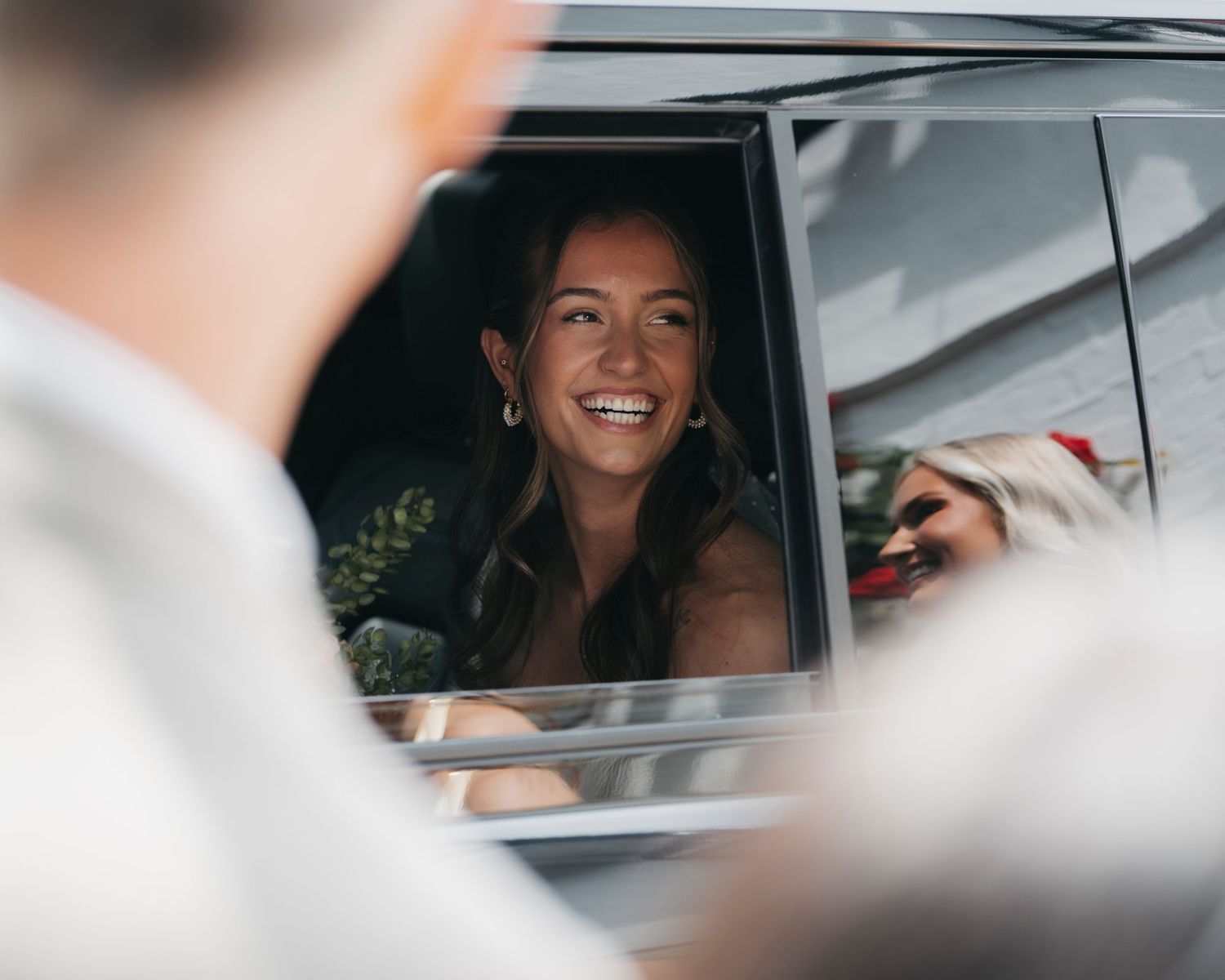Smiling woman with long dark hair looks out from a car window, with a blurred wedding guest in the foreground.