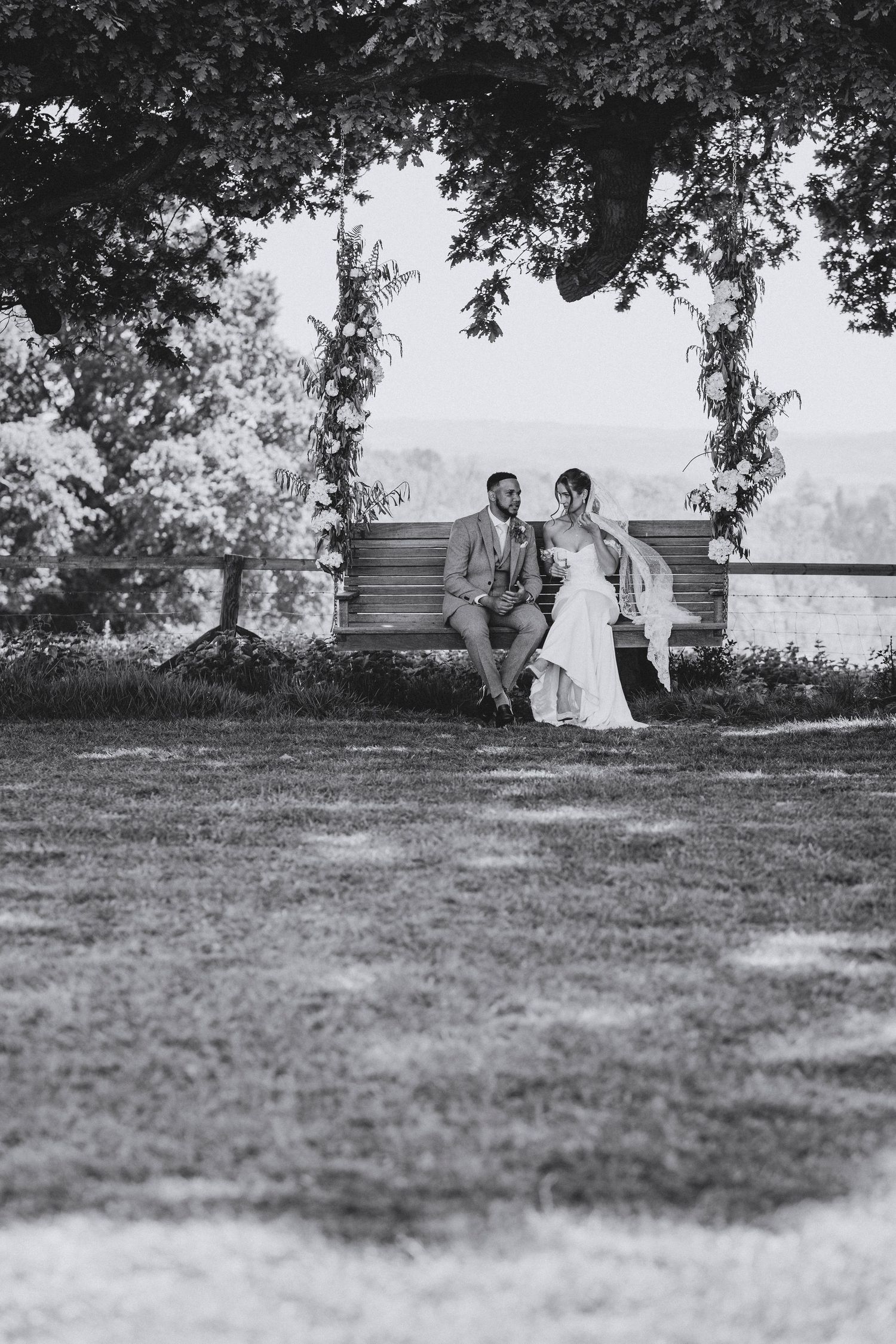 Black and white photo of a bride and groom sitting close on a wooden bench beneath a tree with a flower-covered arch.