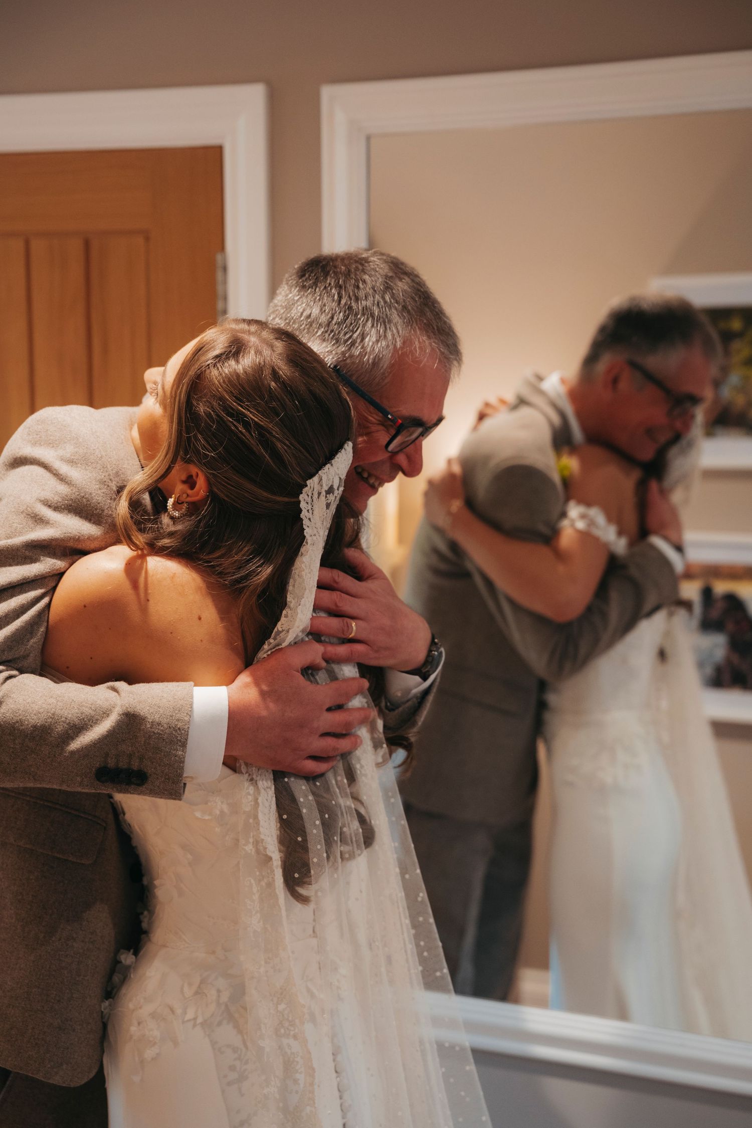 Bride in a lace veil hugs her father tightly in an emotional moment, with their reflection visible in a mirror.