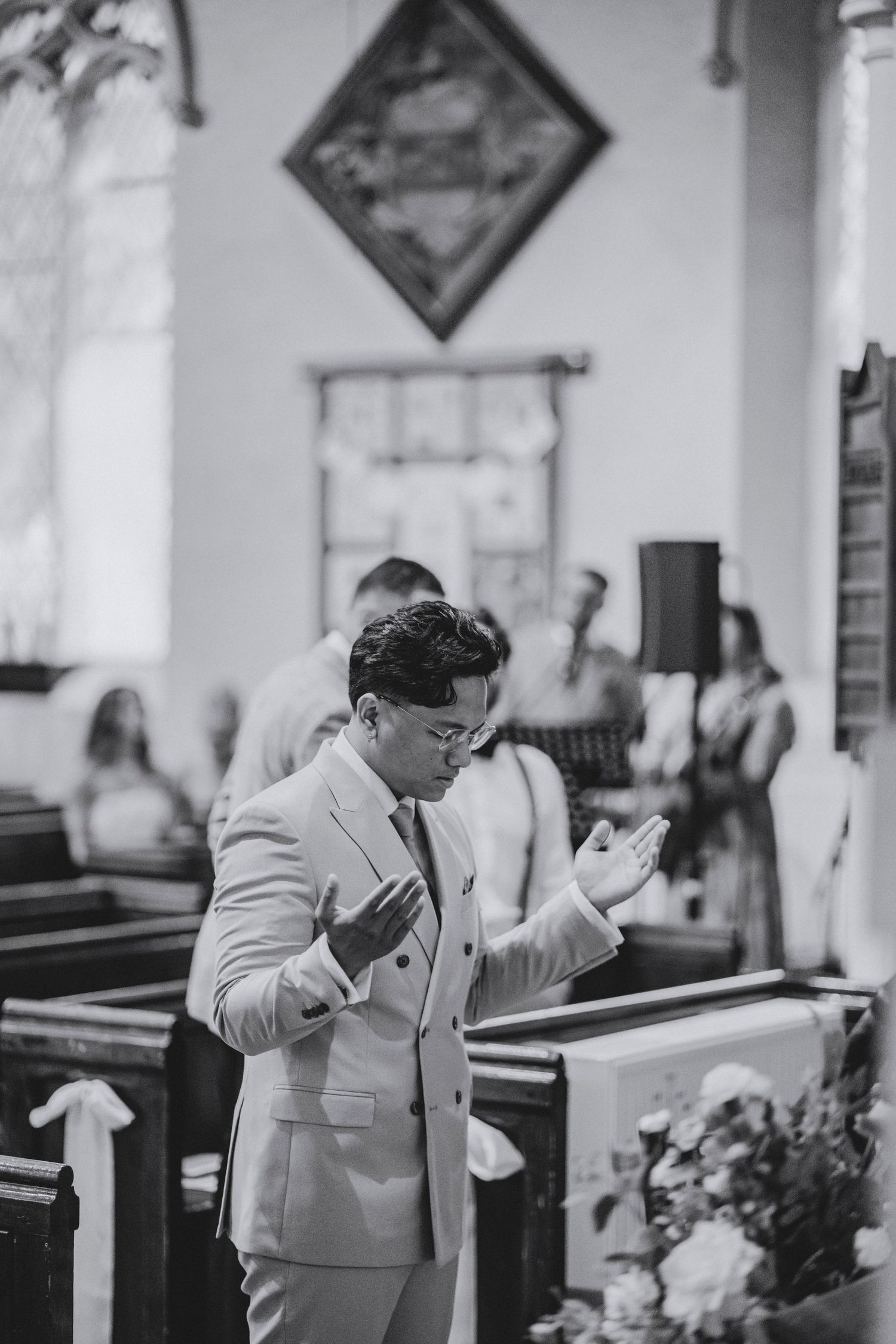 A groom in a light suit raises his open hands in prayer inside a church during a wedding ceremony.