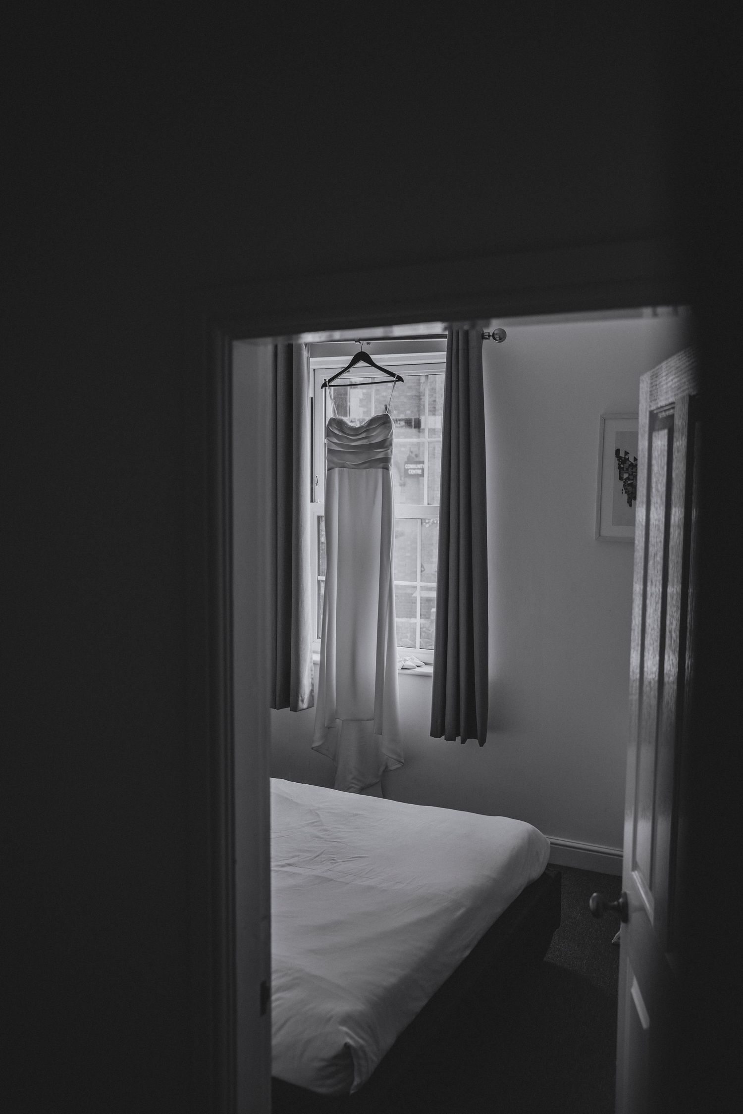 Black and white photo of a wedding dress hanging by a bedroom window, seen through a dark doorway beside a made bed.