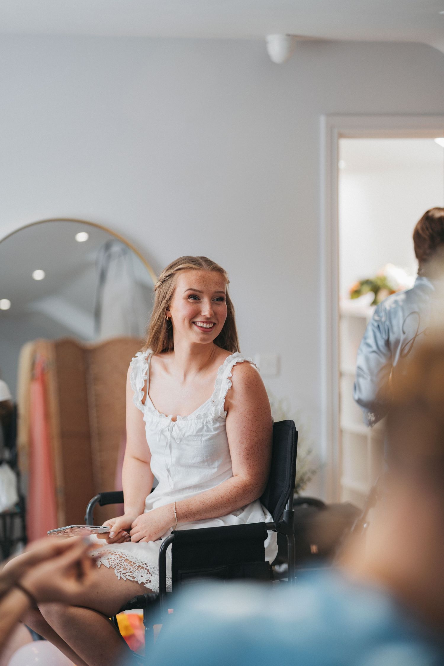 Smiling woman in a white dress sits in a chair during bridal preparations in a bright room with an arched mirror.