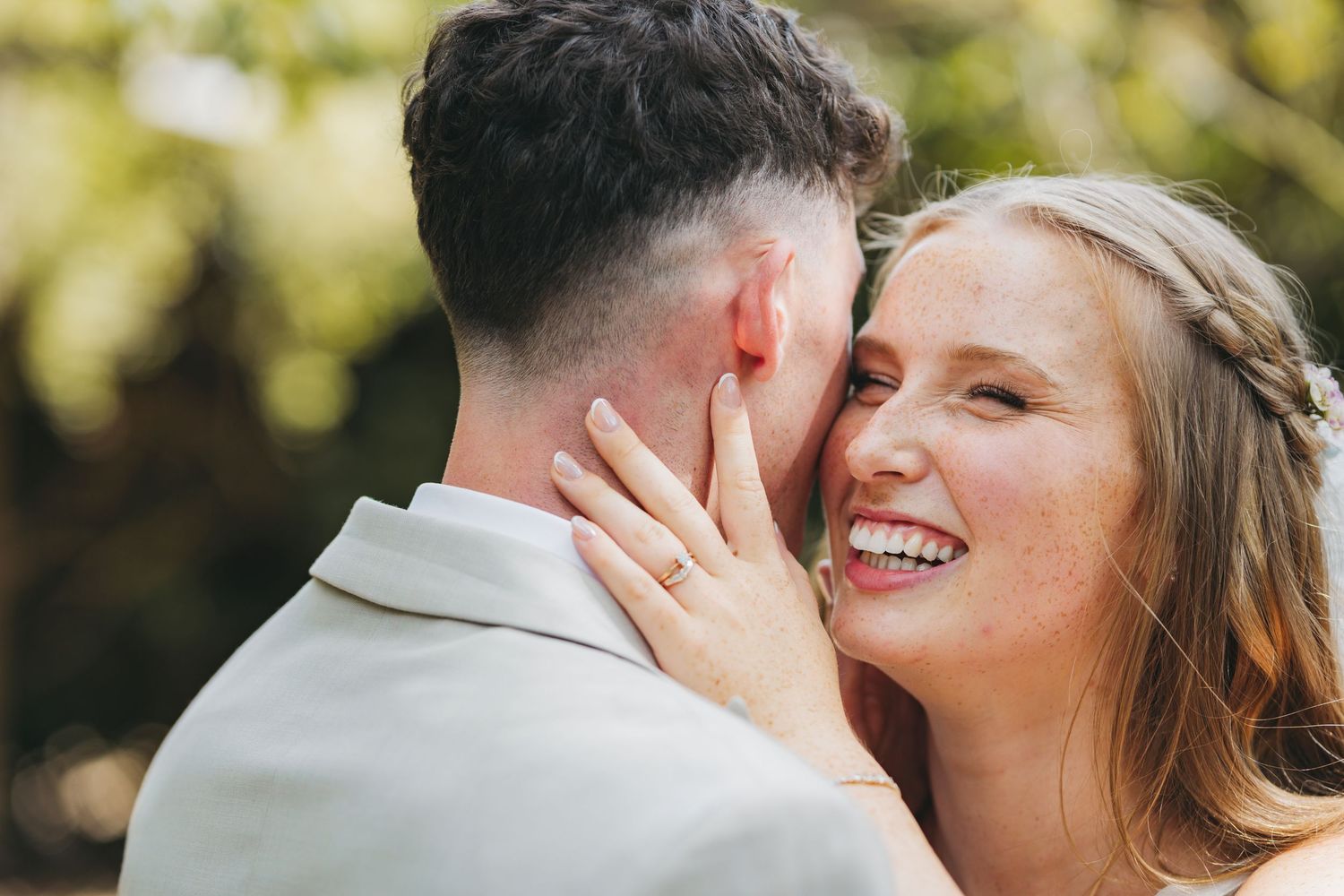 A radiant bride with a braided hairstyle smiles broadly while touching her groom's face in a close wedding portrait.