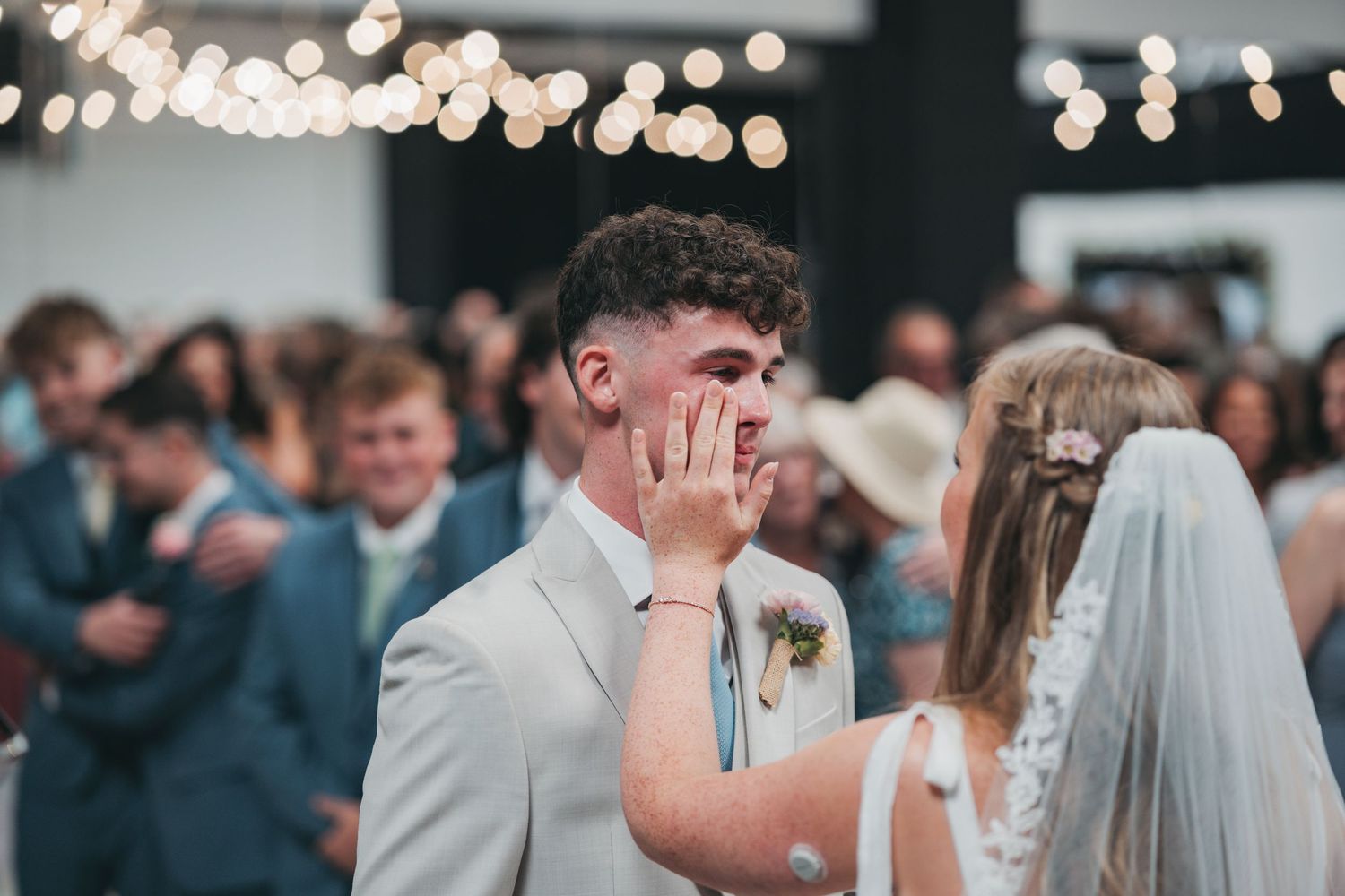 Bride in a veil wipes a happy tear from the groom's face as wedding guests gather behind them under string lights.