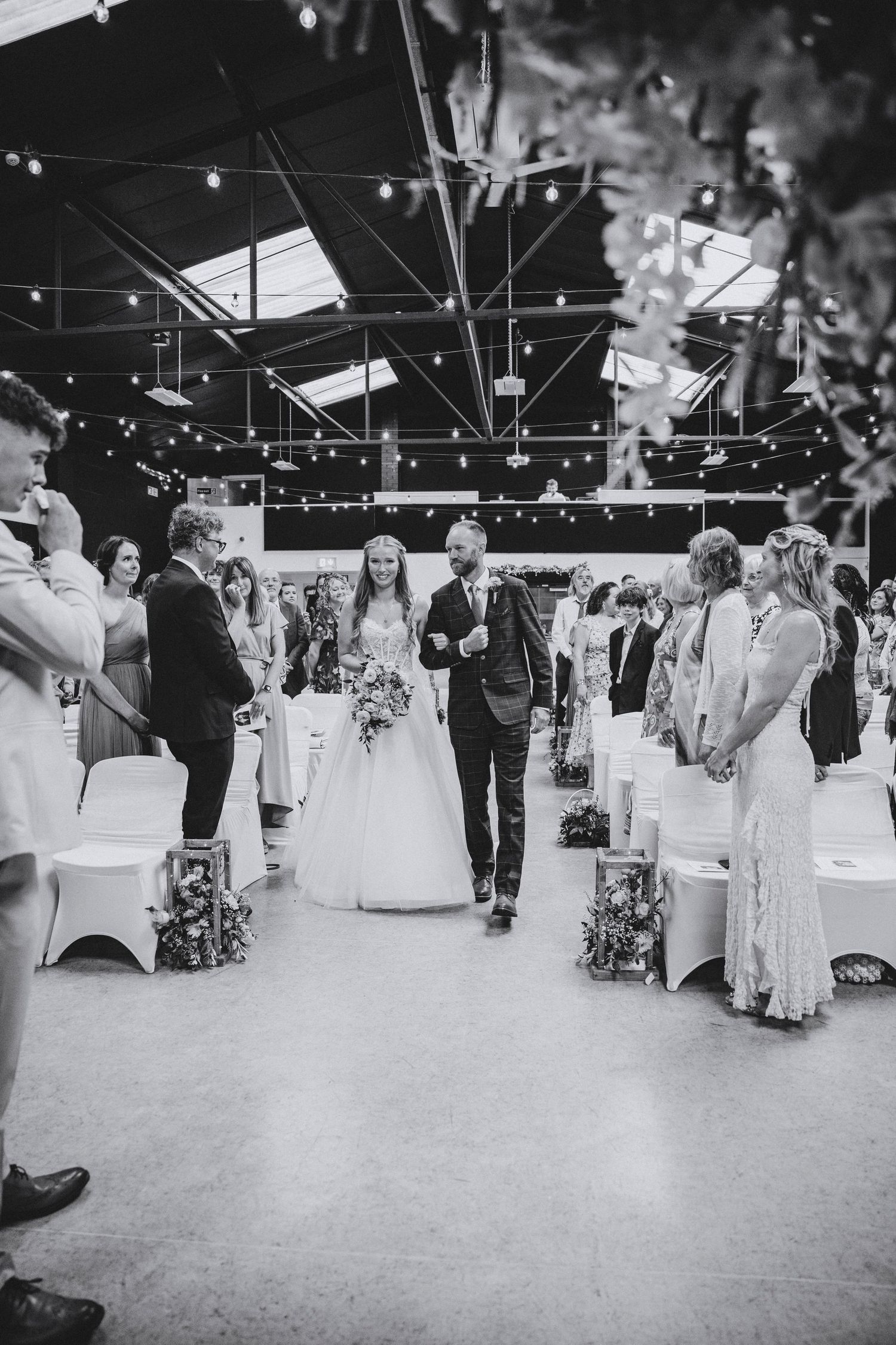 Black and white photo of a father walking the bride in a long gown down the aisle as guests stand and smile.