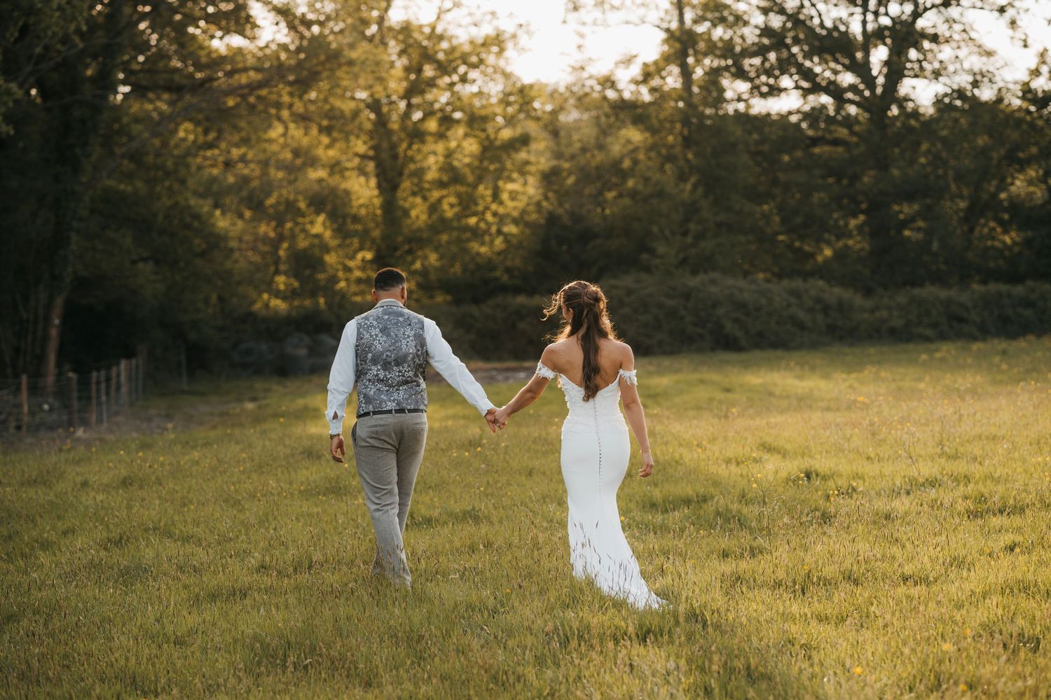 Bride and groom hold hands and walk across a sunlit grassy field at golden hour during their wedding.