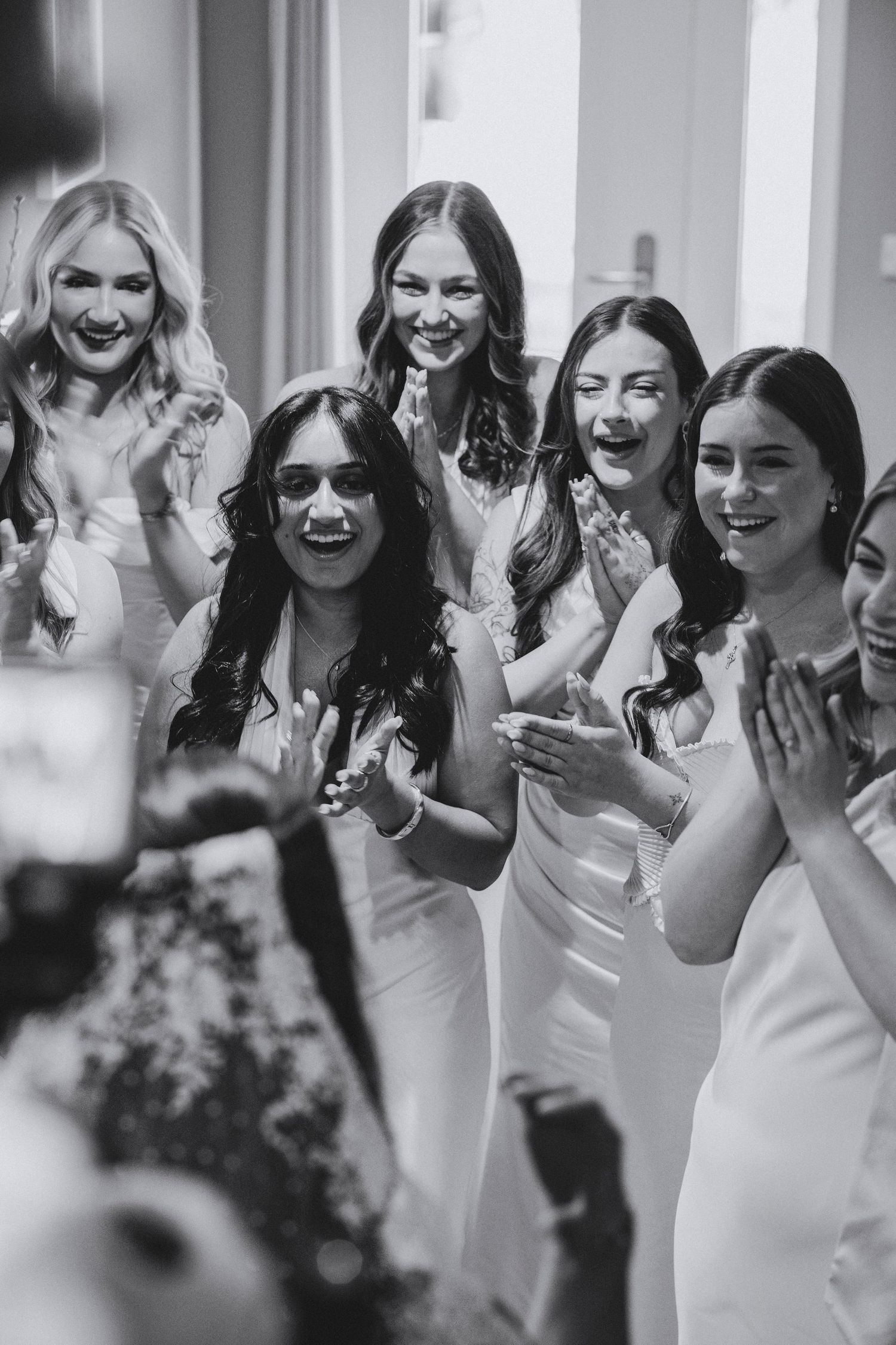 Cheerful bridesmaids in white dresses laugh and clap together in a joyful black and white wedding moment.