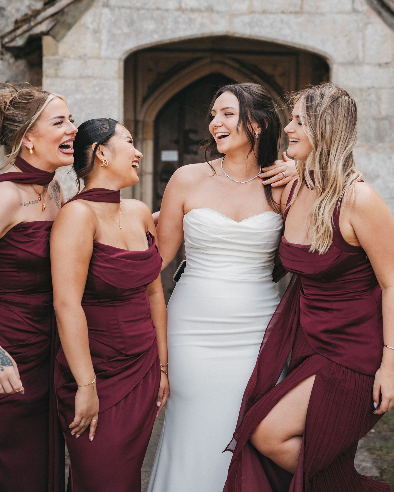 Bride in a white dress laughs with three bridesmaids in burgundy gowns outside a stone church entrance.
