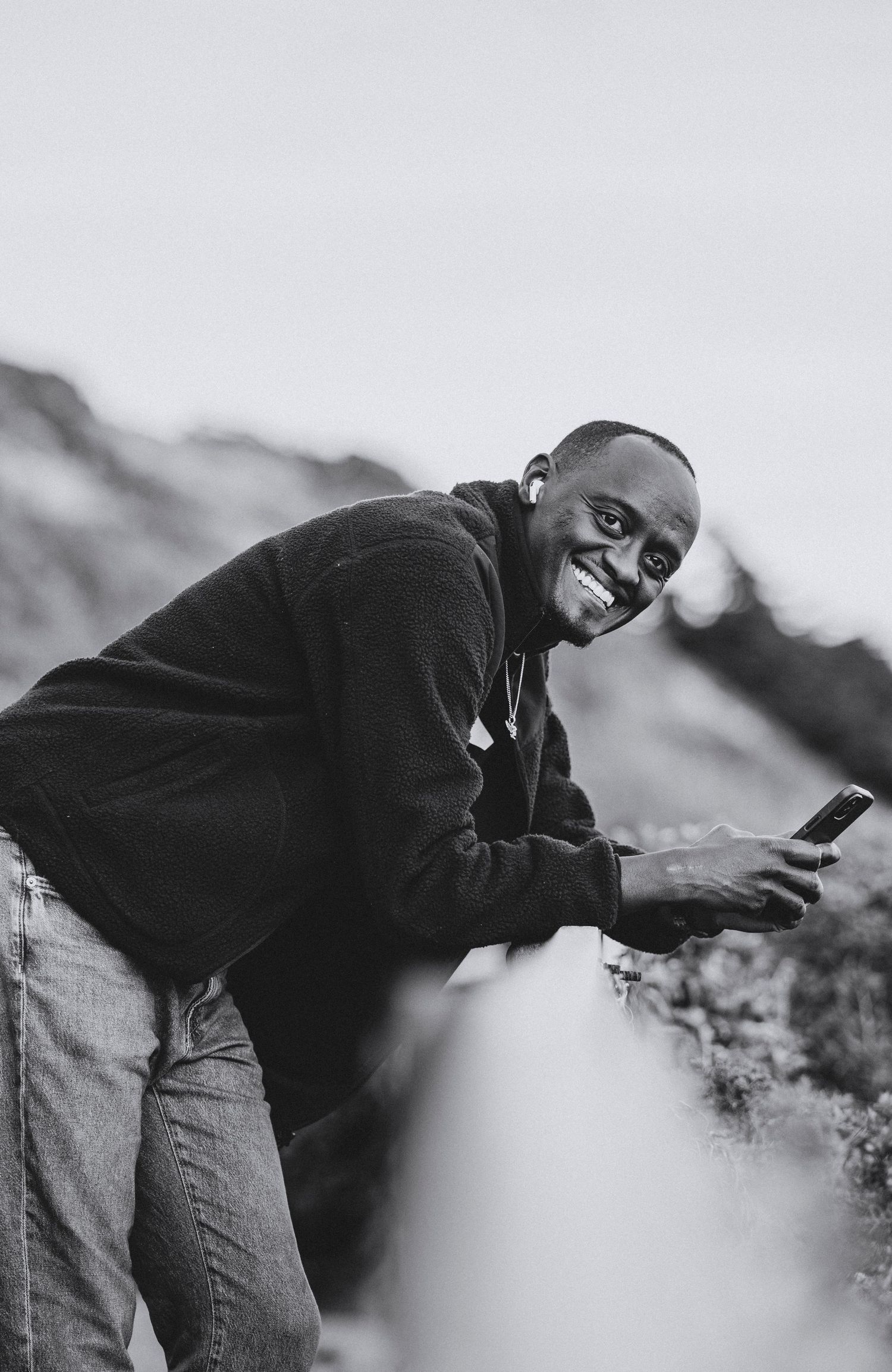 Black and white portrait of a smiling man leaning on a railing while holding his phone, wearing earbuds and a fleece jacket.